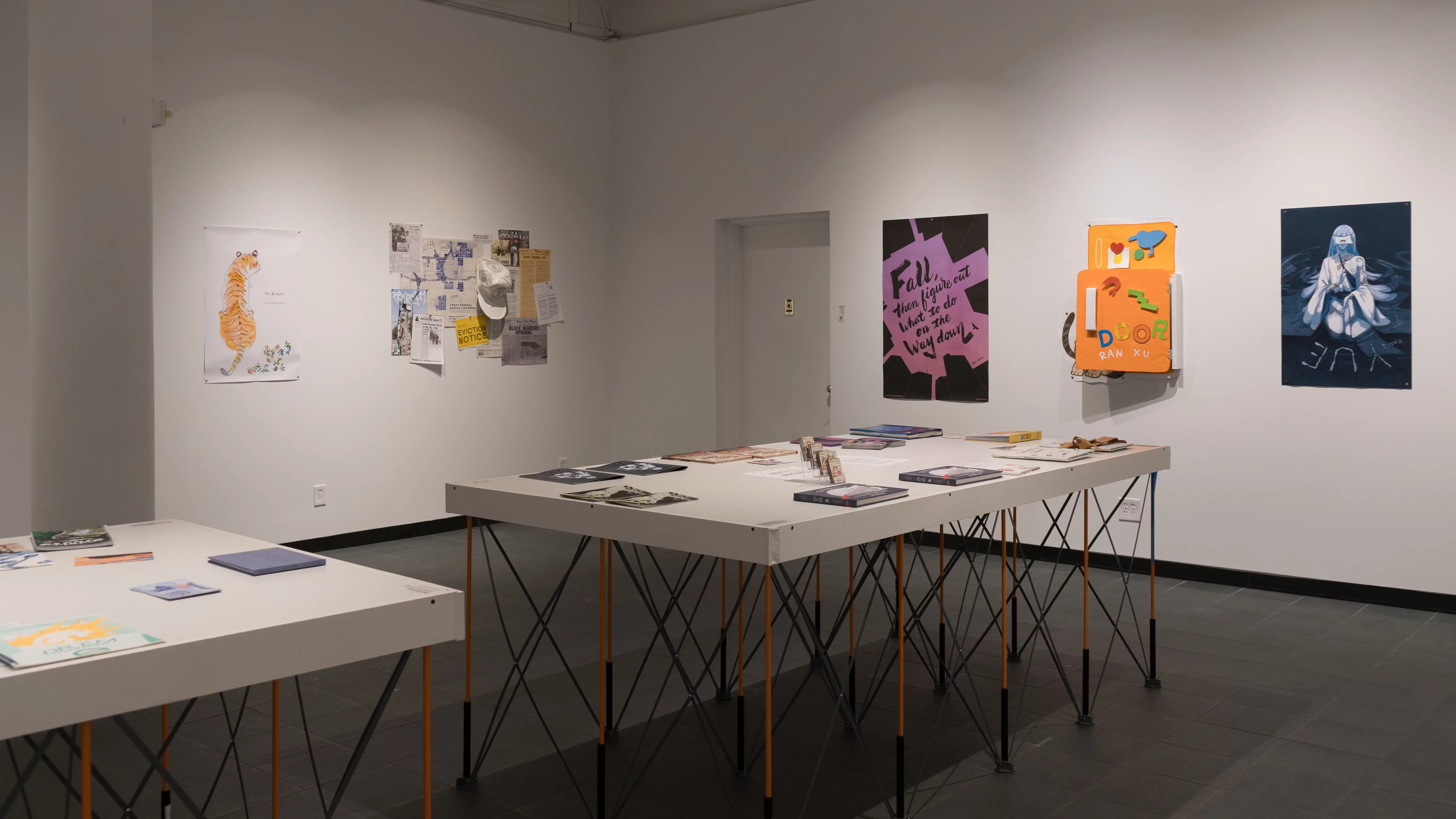 Two tables in the gallery display many books, laid flat. Prints, posters, layered papers, and cardboard sculptures are hung on the surrounding walls. The works vary in color, size, style, and content.