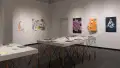 Two tables in the gallery display many books, laid flat. Prints, posters, layered papers, and cardboard sculptures are hung on the surrounding walls. The works vary in color, size, style, and content.