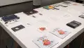 Press releases and a gallery map lay in the center of a table in the gallery. Many different books are arranged around the papers. The nearest book is titled “Bumbling Bud” and depicts a smiling red and orange bumblebee-like creature.
