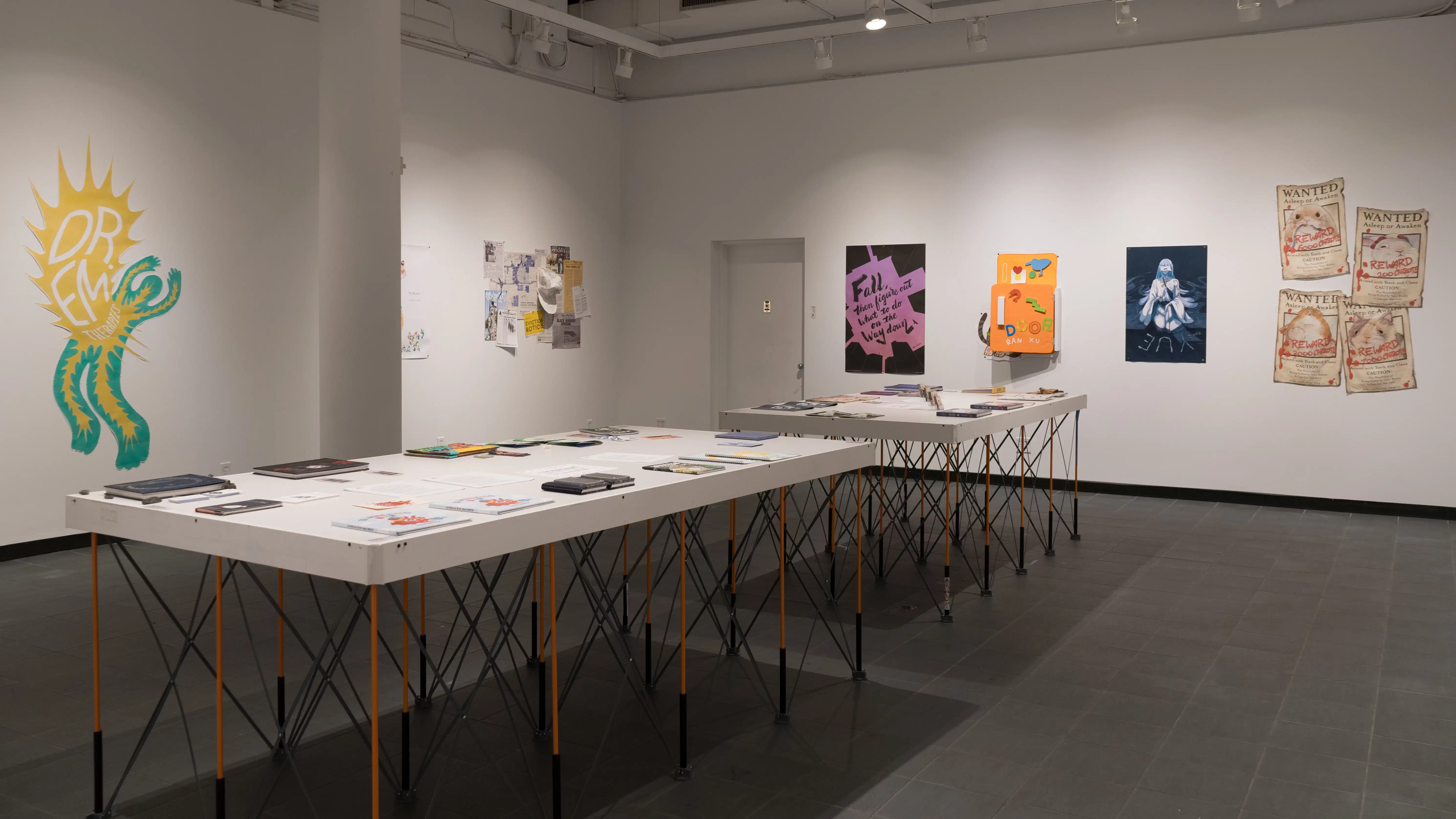 Looking towards one corner of the gallery, two tables, covered with various books, are fully visible. Different types of illustrated works are displayed on the surrounding walls, including prints, sculptures, and installations made from layered paper.