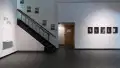 A wall in SVA’s Gramercy Gallery, with a staircase on the left side. Beside it is the name of the exhibition, and to the right are a line of photographs.