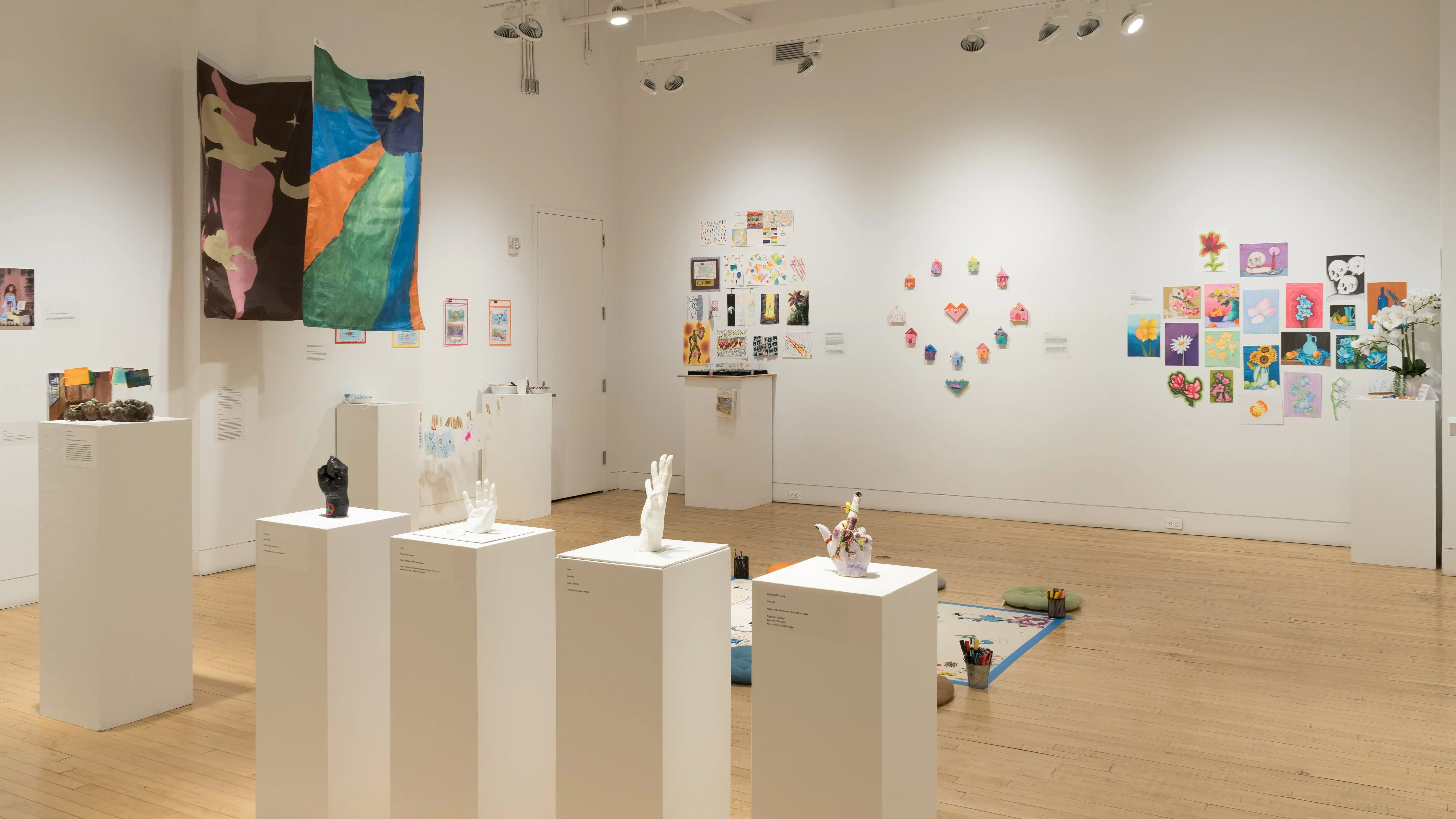 Inside the SVA Flatiron Gallery, works in various mediums, sizes, and colors cover the walls and rest on the floor or on pedestals. Sculptures of human hands sit on a row of pedestals near the front of the gallery.