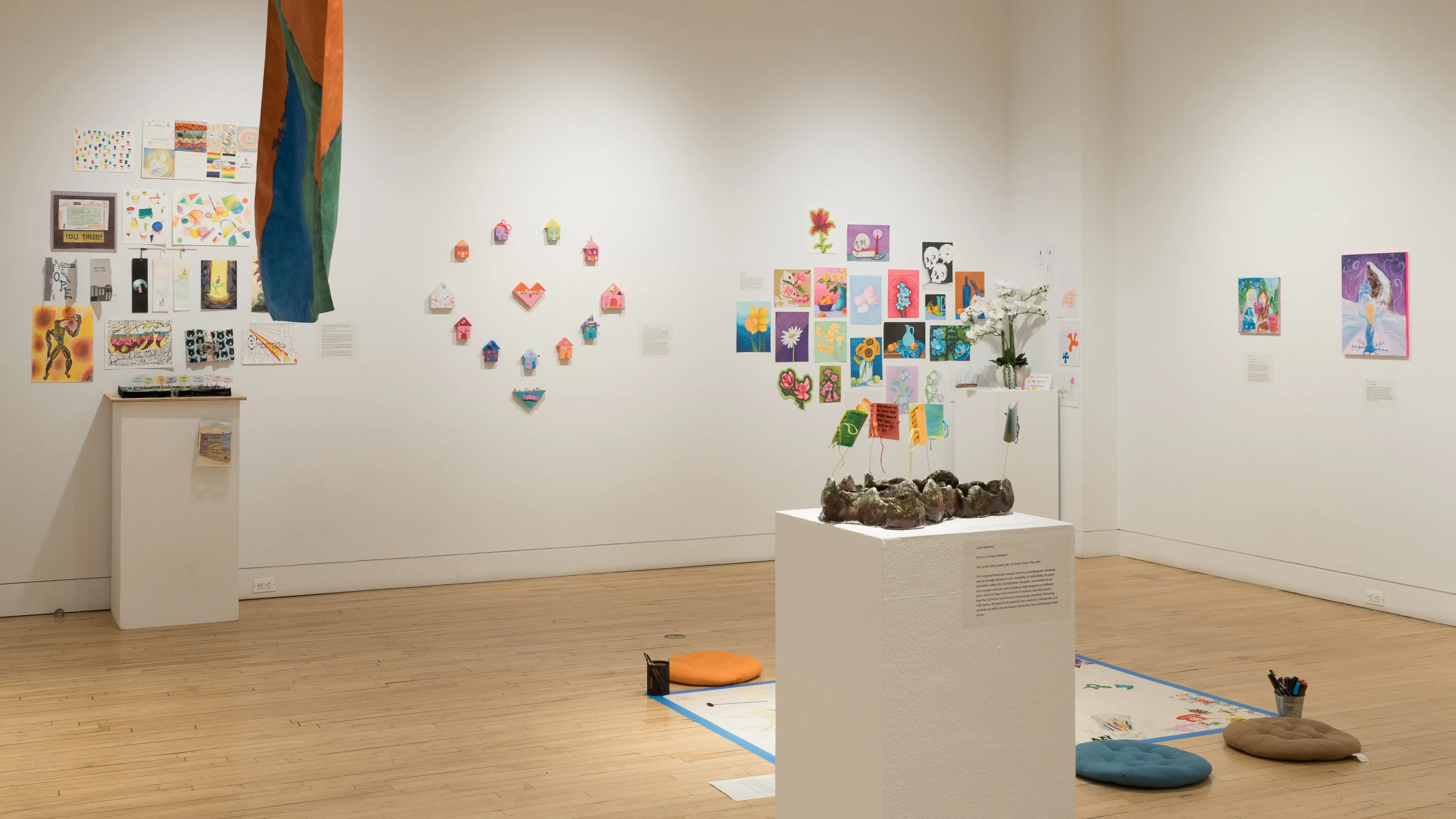 In the center of a wall, small objects shaped like hearts and houses are arranged in a circle. In groups on the left and right of the installation, many drawings and paintings on paper are hung close together. In the foreground, a brown ceramic sculpture rests on a pedestal.