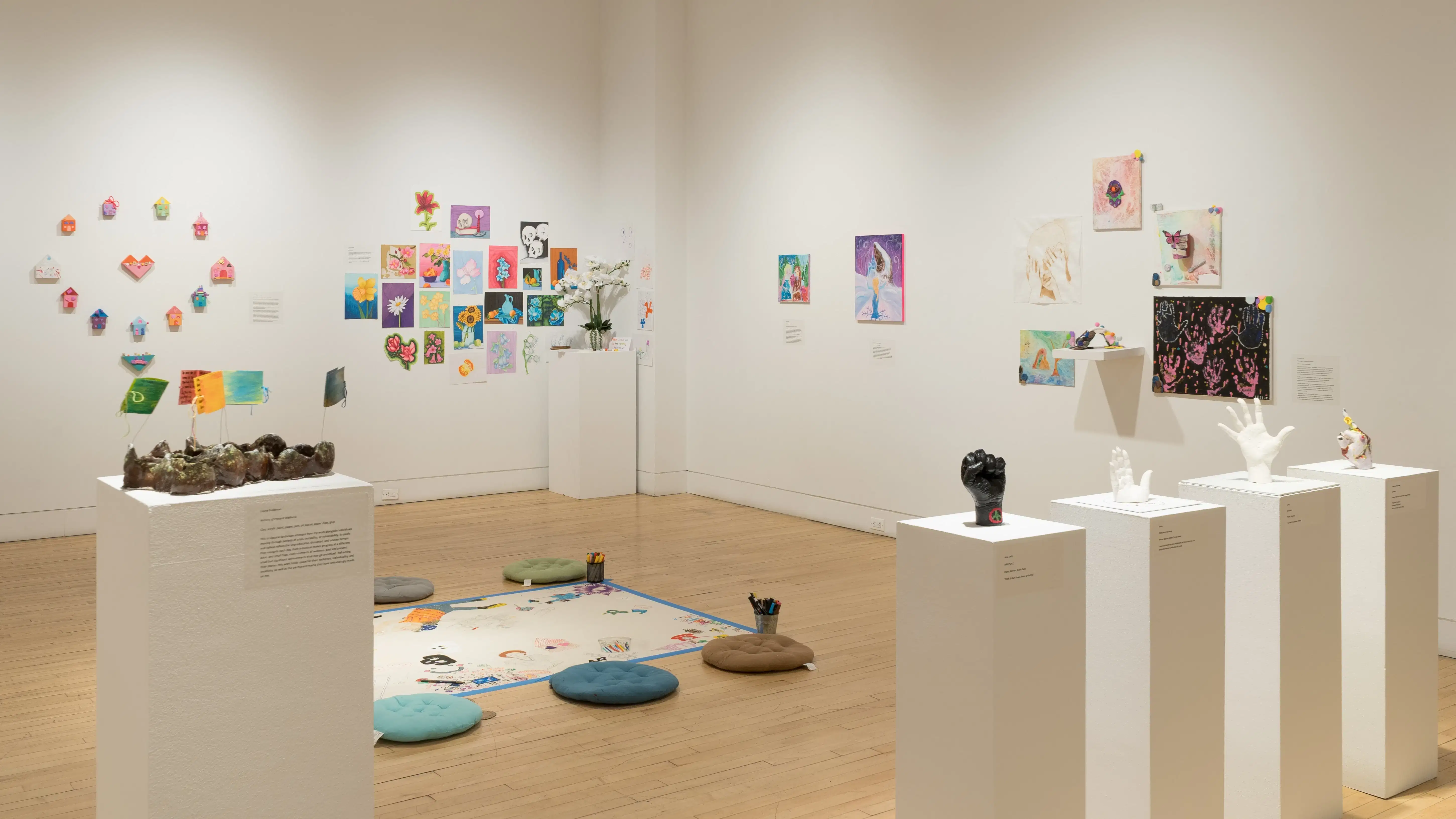 In the center of the room, a large posterboard is taped to the floor and is surrounded by floor cushions, inviting visitors to draw on the board. Many works hang on the surrounding walls, including paintings on canvas, drawings on paper, and small sculptures. A row of pedestals holds sculptures of human hands.