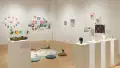 In the center of the room, a large posterboard is taped to the floor and is surrounded by floor cushions, inviting visitors to draw on the board. Many works hang on the surrounding walls, including paintings on canvas, drawings on paper, and small sculptures. A row of pedestals holds sculptures of human hands.