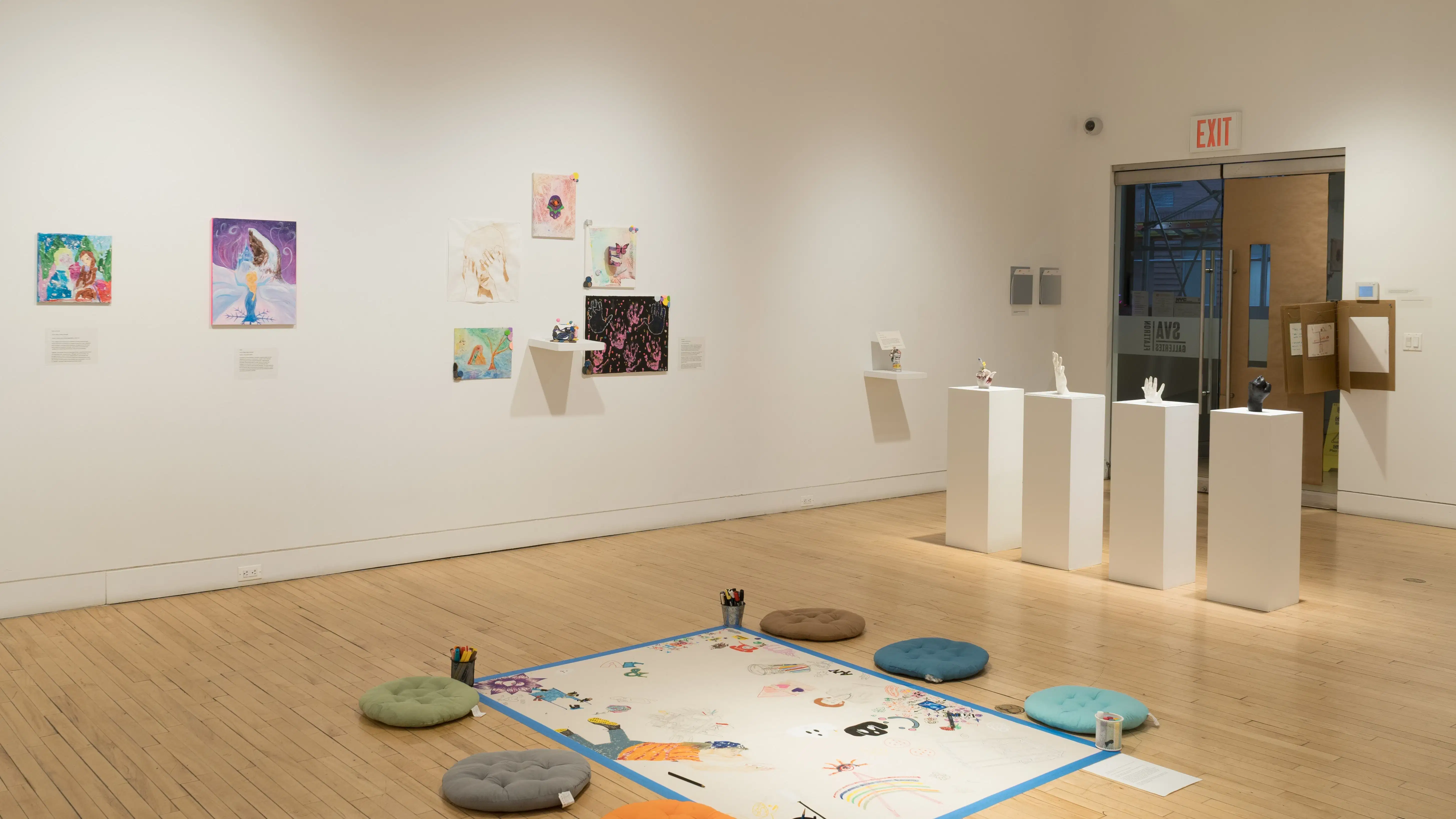 In the center of the room, a large posterboard is taped to the floor and is surrounded by floor cushions, inviting visitors to draw on the board. In front of them, four sculptures of human hands are displayed on pedestals. Canvas and paper works hang on the wall beside them.