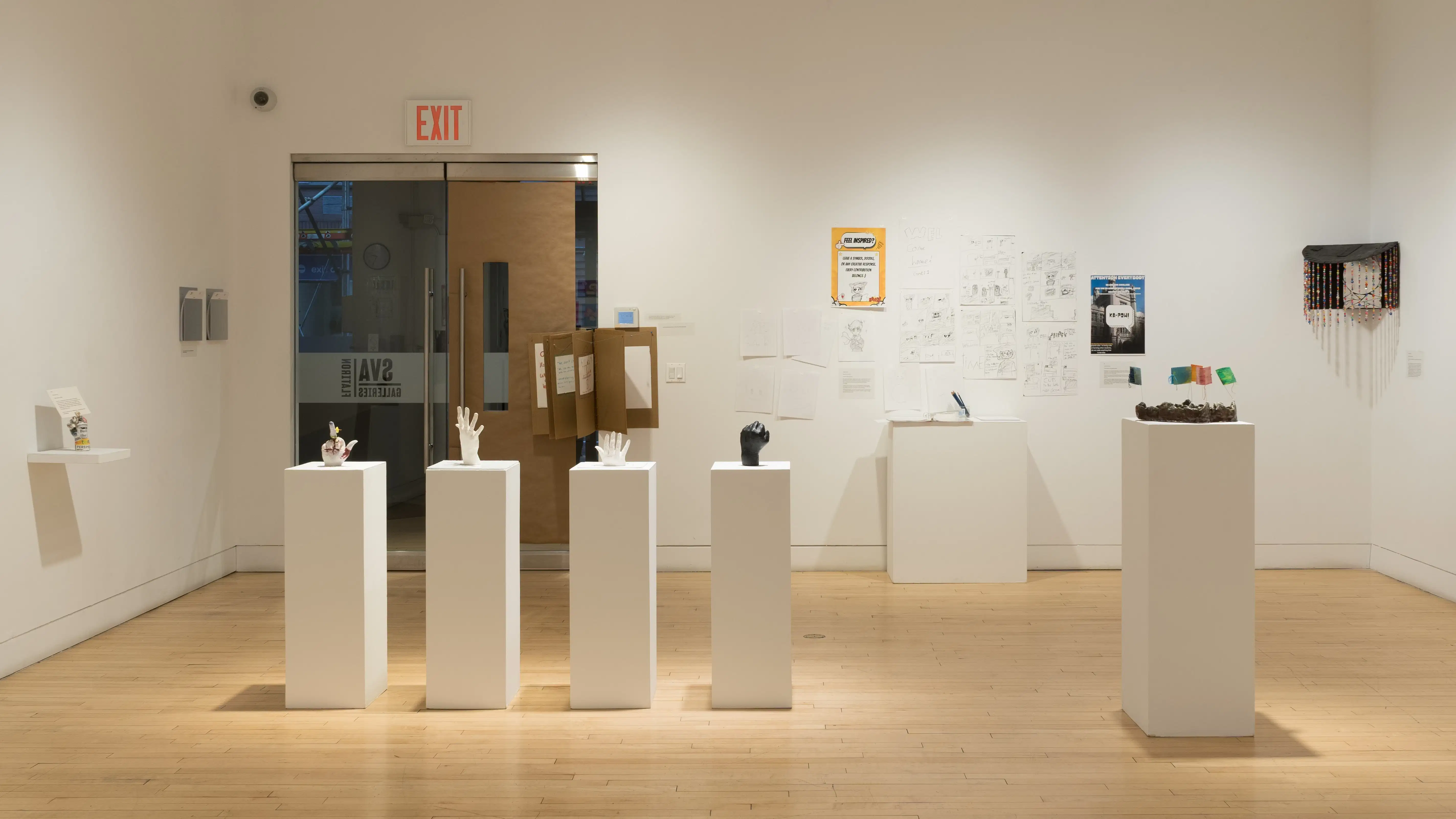 A row of pedestals sits almost in front of the glass gallery doors. To the right of the doors, several pencil drawings on paper are displayed above another pedestal.