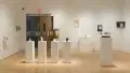 A row of pedestals sits almost in front of the glass gallery doors. To the right of the doors, several pencil drawings on paper are displayed above another pedestal.