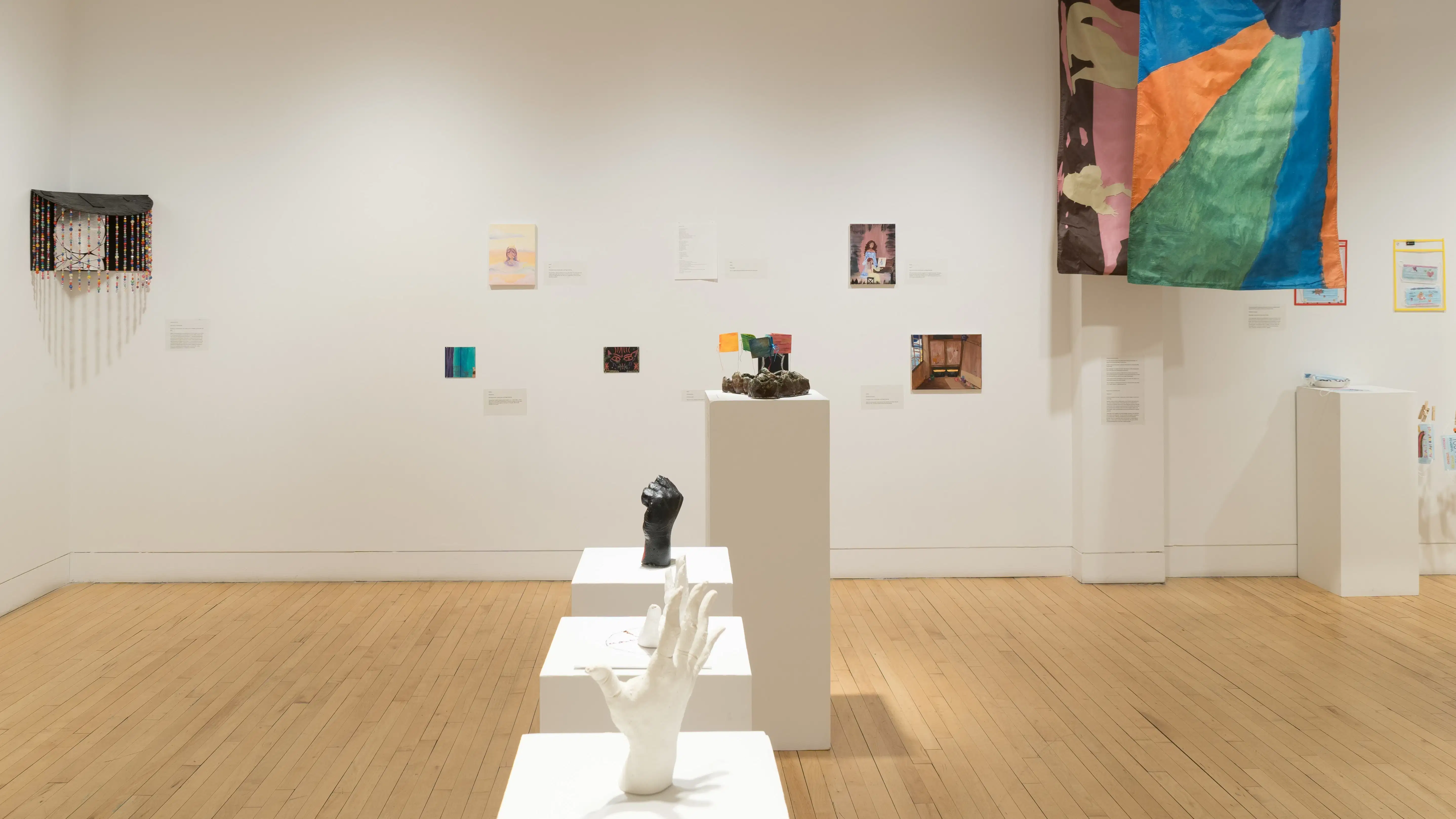 A black box-type sculpture with a beaded curtain hangs in the corner between two walls. To the right, several small paintings hang to the left of two suspended flags.