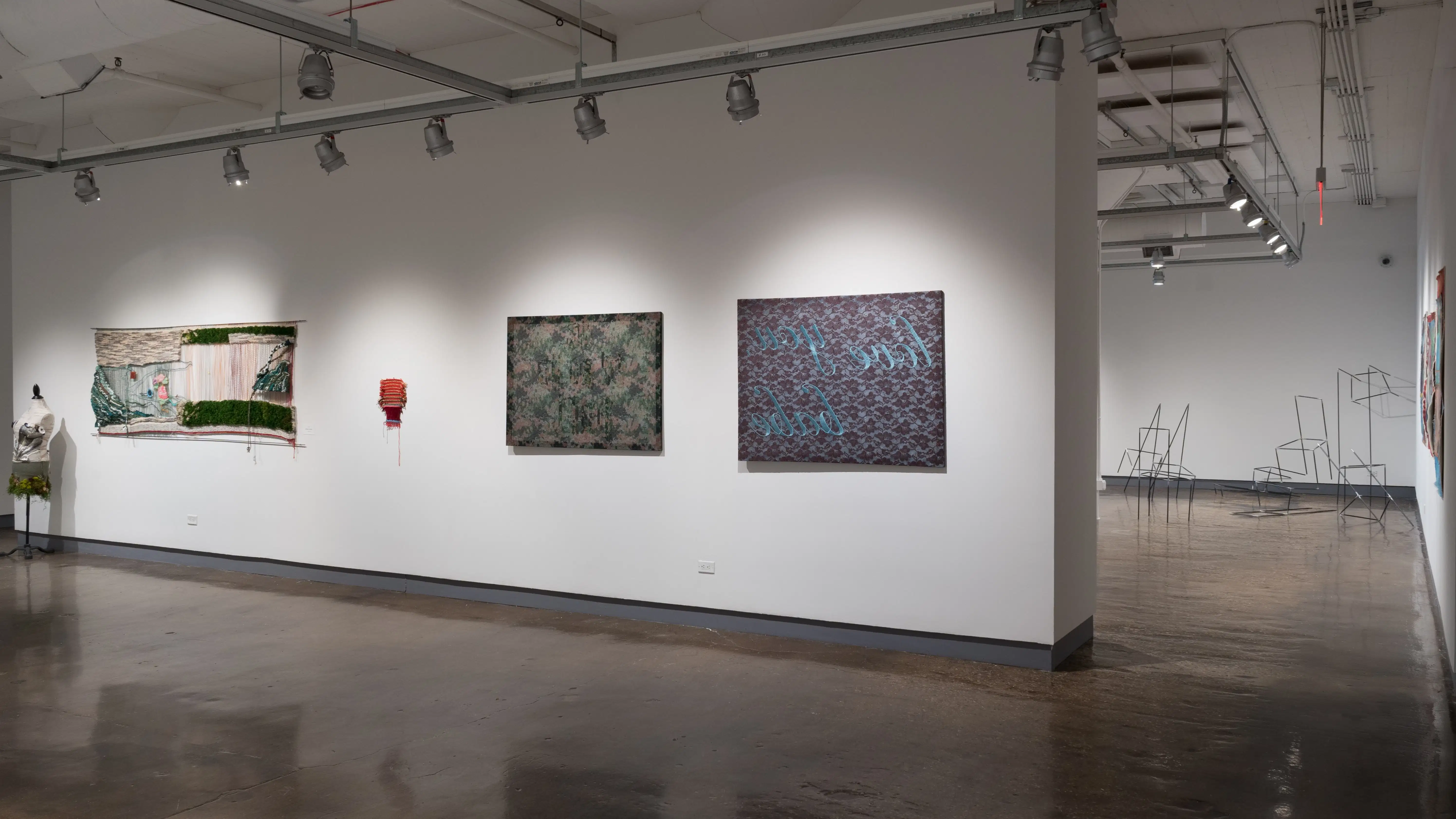 On the wall in the second gallery looking into the first gallery, mixed media work hangs on the wall, with sculptural work in the background.