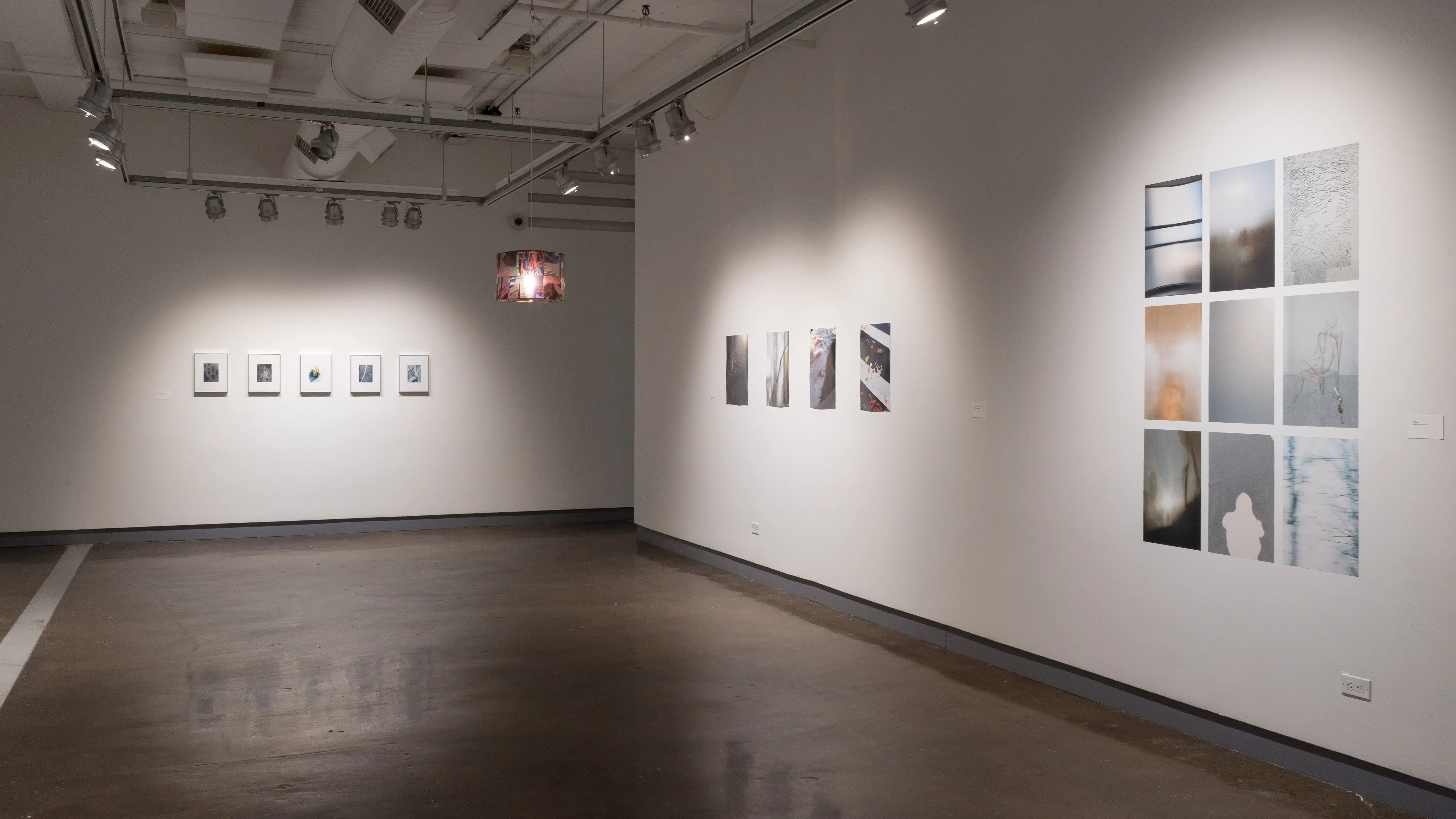 Five small, matted and framed photographs hang in a row at the far end of gallery two. A colorful lamp is suspended from the ceiling. Four printed photographs are pinned side by side on the right wall, and further down, nine photographs are arranged in a 3 x 3 grid.