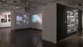From the right hallway, most of the alcove in gallery three is visible. Inside the alcove, images of people’s faces, cropped around the eyes, are projected onto the walls. A portrait on fabric is suspended beside them. Four large photographs of a concert are visible on the farthest wall across the room. In the right hallway, many black and white photographs are grouped together in a loose grid.