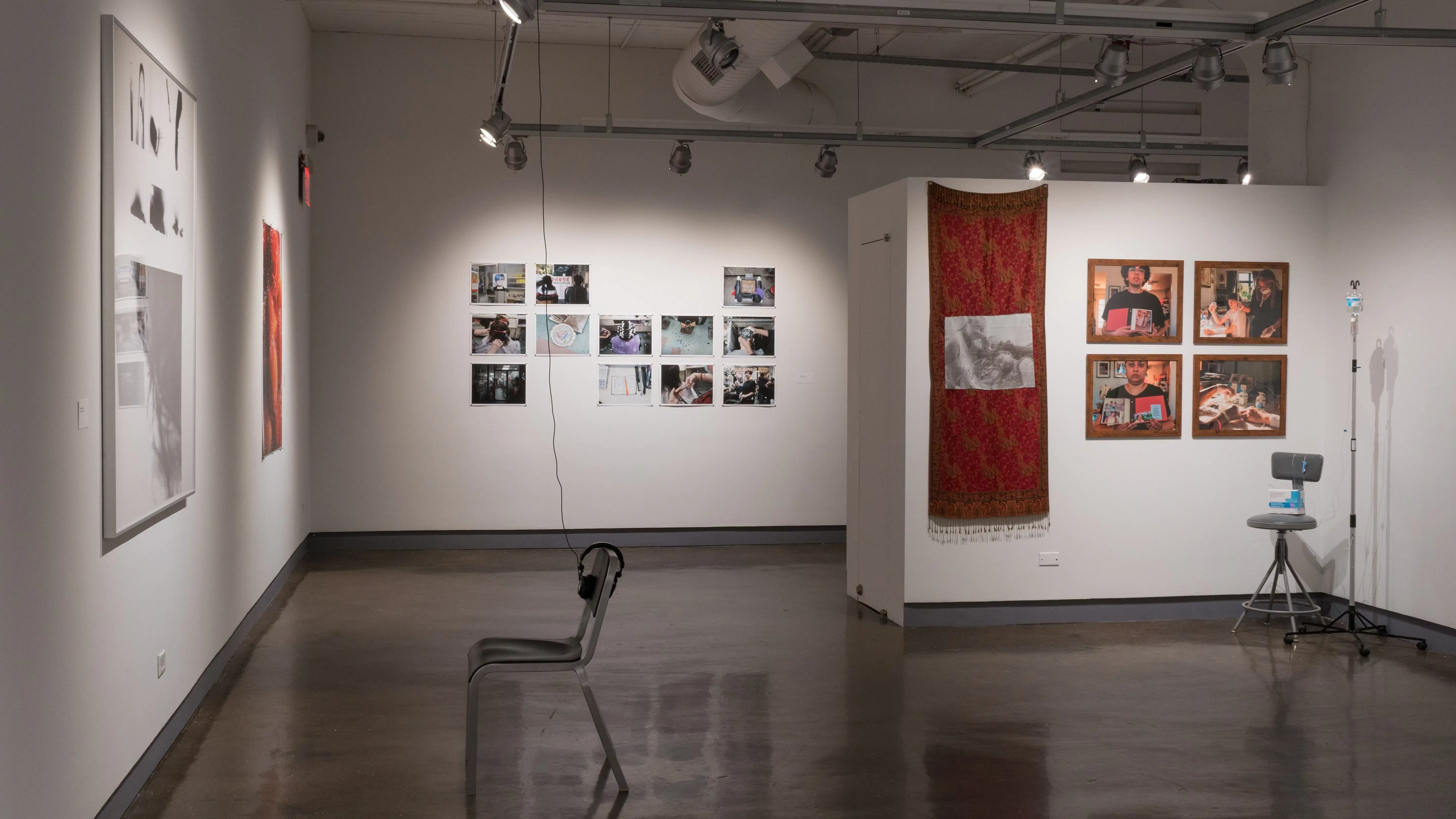 On the far wall of gallery four, many photographs are arranged in a grid, with some slots left empty. The parallel wall inside the alcove displays four medium-sized framed photographs next to a long red scarf. A doctor’s office chair and an IV drip stand are set up beside them. Two large, separate prints hang on the perpendicular wall, with a chair and headphones set up to face them.