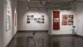On the far wall of gallery four, many photographs are arranged in a grid, with some slots left empty. The parallel wall inside the alcove displays four medium-sized framed photographs next to a long red scarf. A doctor’s office chair and an IV drip stand are set up beside them. Two large, separate prints hang on the perpendicular wall, with a chair and headphones set up to face them.