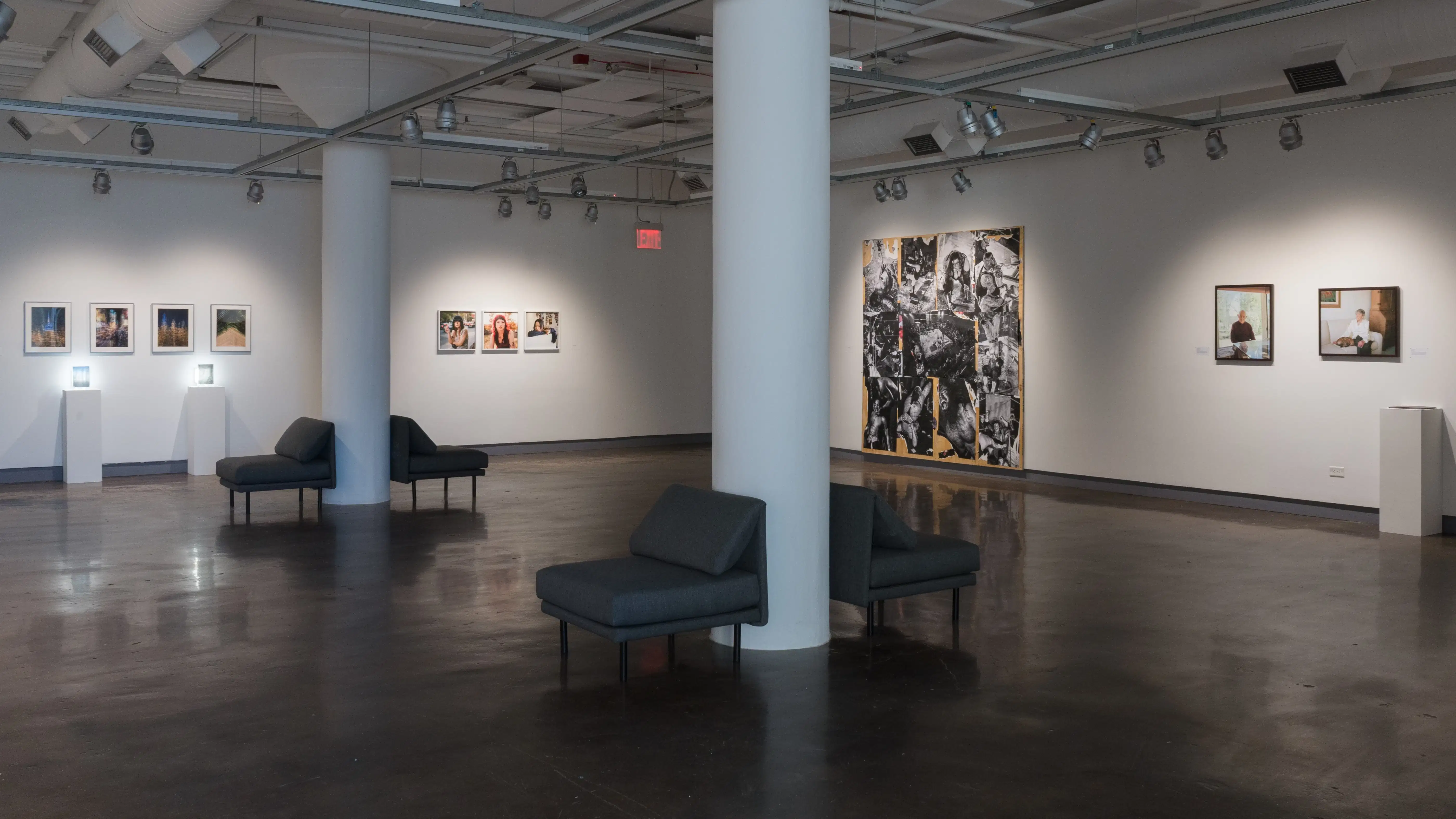 A large piece of plywood covered in black and white photographs is attached to a wall in gallery one. To the right and left of it, medium-sized framed photographs hang in a row. On the far left, two lights glow behind small works on pedestals.