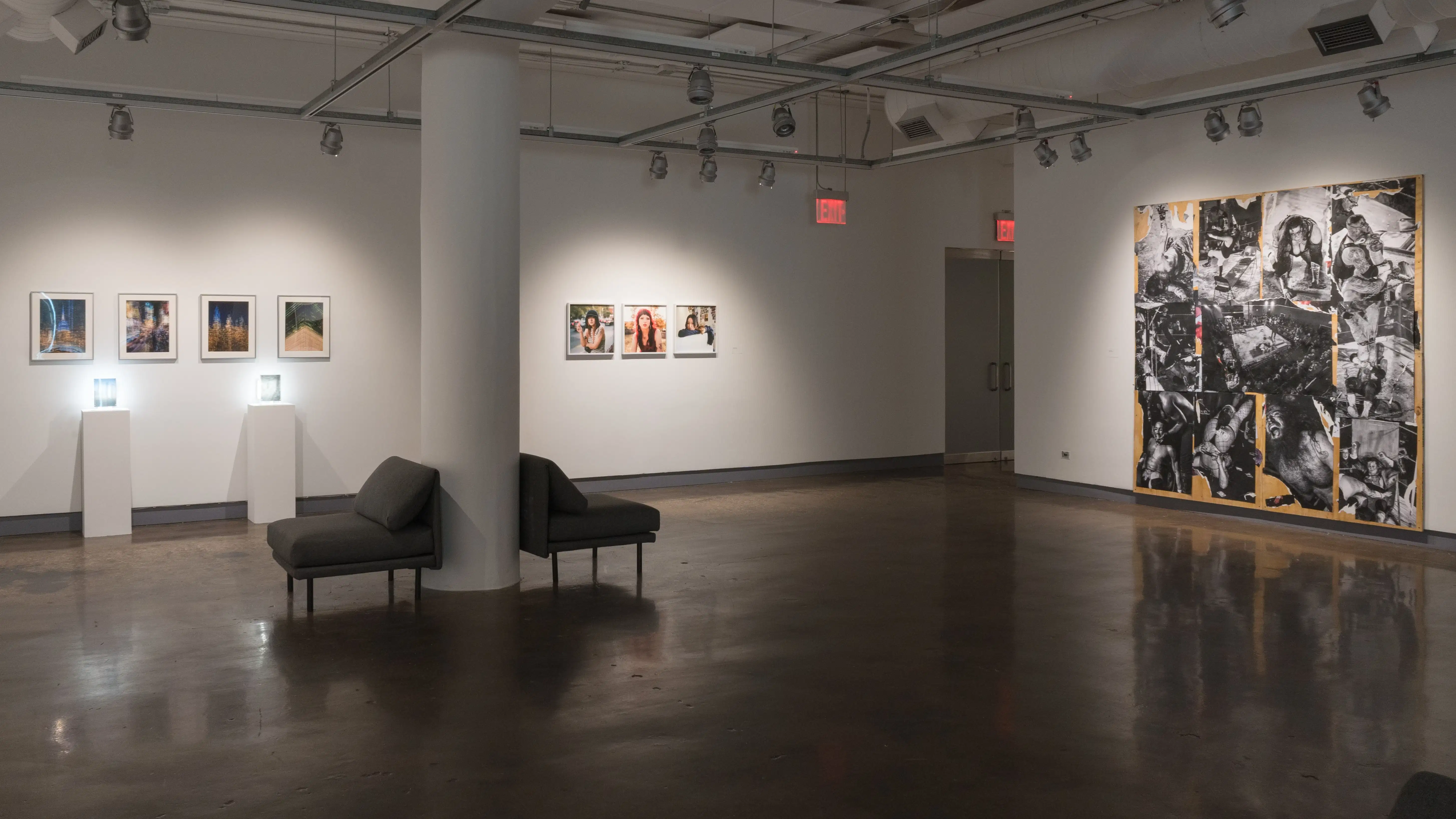 A large piece of plywood covered in black and white photographs is attached to a wall in gallery one. On the far left, two lights glow behind small works on pedestals, underneath a row of framed photographs. Between these two sections, three medium-sized framed photographs hang side by side.