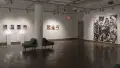 A large piece of plywood covered in black and white photographs is attached to a wall in gallery one. On the far left, two lights glow behind small works on pedestals, underneath a row of framed photographs. Between these two sections, three medium-sized framed photographs hang side by side.