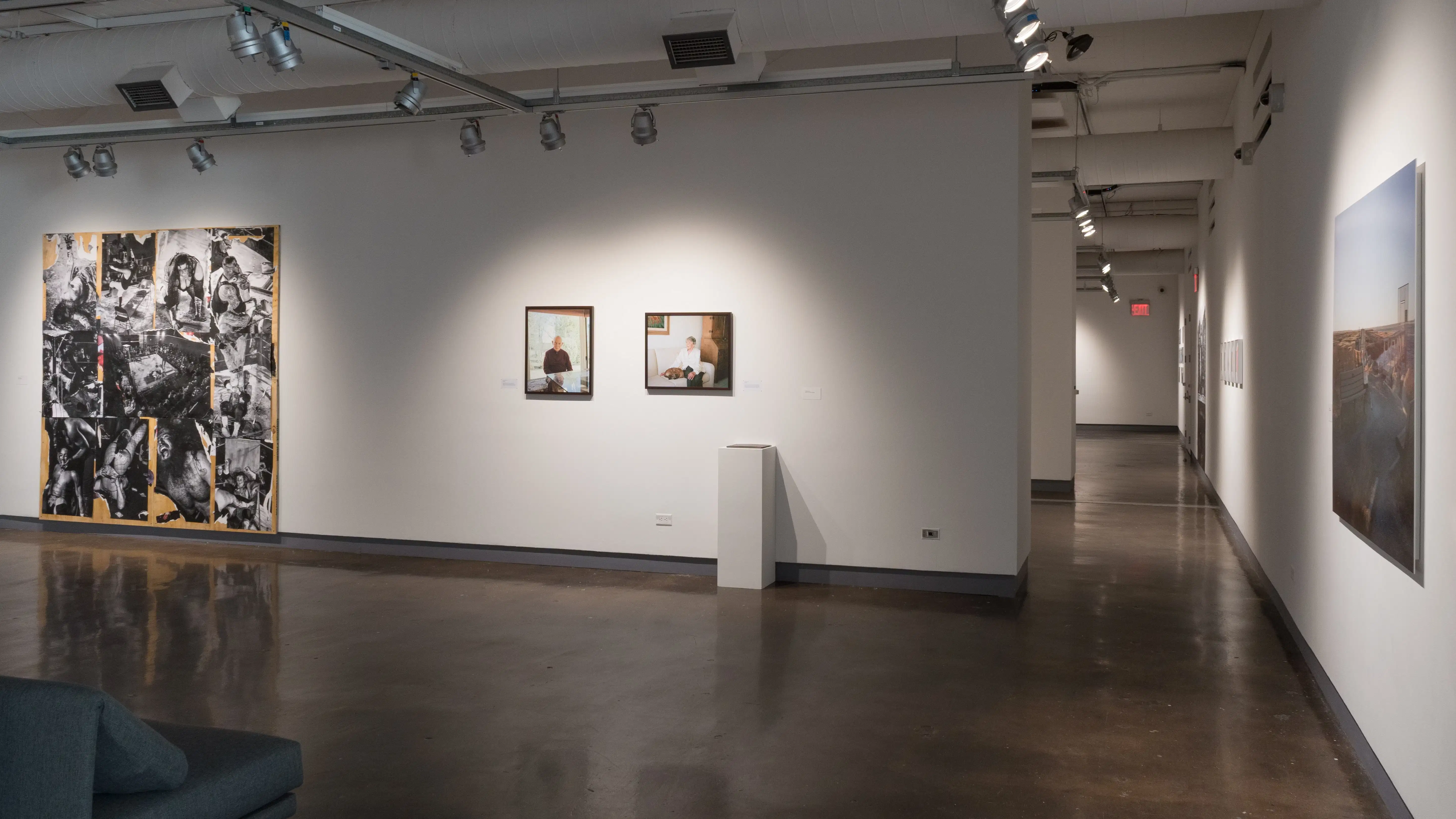 In a corner of gallery one, a large, single photograph is hung in the center of the right wall. Diagonally across, there is a large piece of plywood covered in black and white photographs attached to the wall. Between these two sections, two framed color photographs hang above a single pedestal.