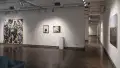 In a corner of gallery one, a large, single photograph is hung in the center of the right wall. Diagonally across, there is a large piece of plywood covered in black and white photographs attached to the wall. Between these two sections, two framed color photographs hang above a single pedestal.