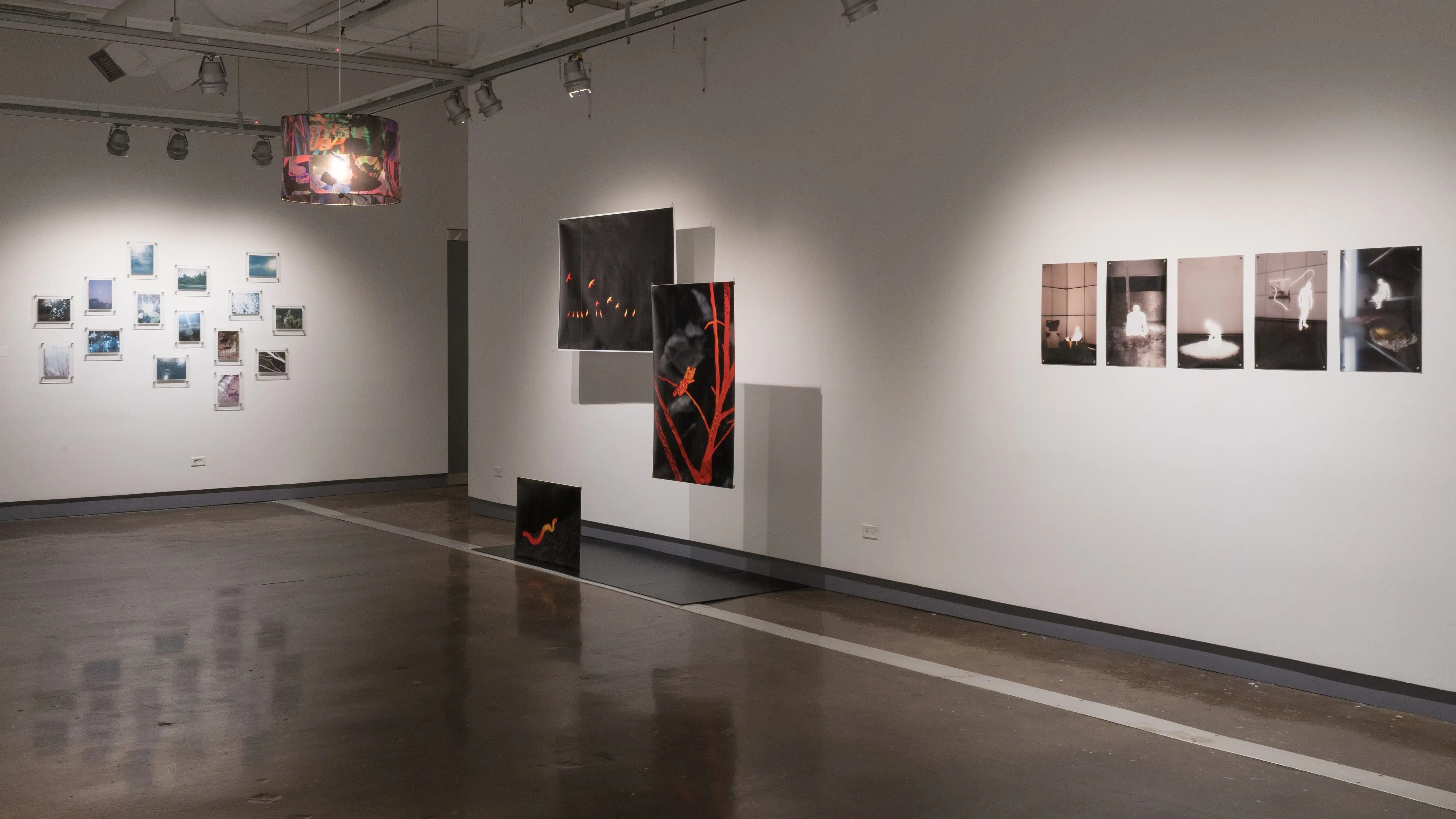Many smaller, mostly cool-toned photographs are displayed in a constellation-like arrangement on the far wall of gallery two. A colorful lamp hangs from the ceiling in the center of the room. Three black and red photographs are suspended in front of the right wall, and further to the right, a row of five images is pinned on the wall.