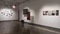 Many smaller, mostly cool-toned photographs are displayed in a constellation-like arrangement on the far wall of gallery two. A colorful lamp hangs from the ceiling in the center of the room. Three black and red photographs are suspended in front of the right wall, and further to the right, a row of five images is pinned on the wall.