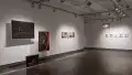 Three black and red photographs are suspended in a group in front of the left wall. Towards the right, a row of five images is pinned on the wall. On the wall perpendicular to it, five small, matted and framed photographs hang in a row. A colorful lamp is suspended from the ceiling.