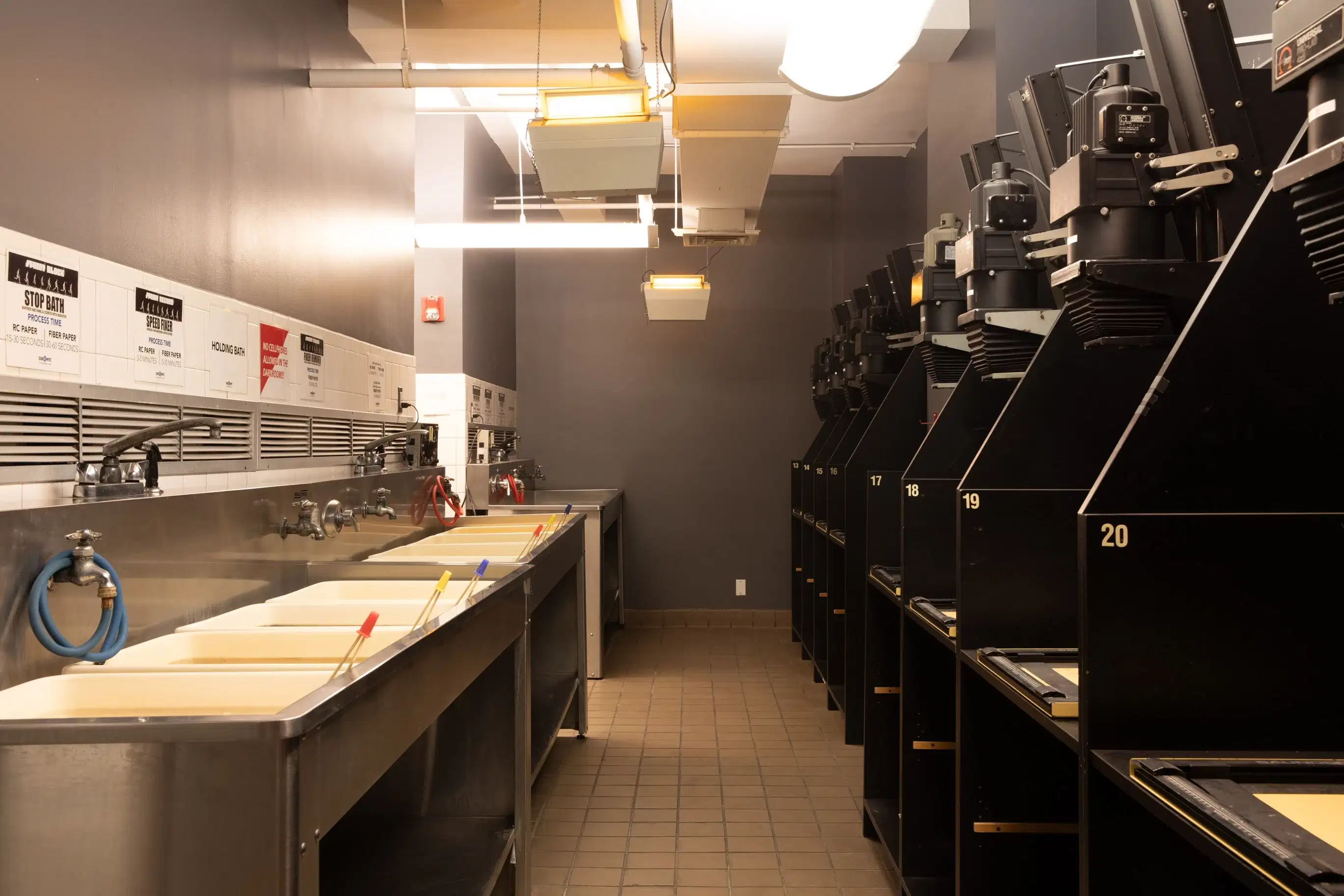 Dark room with a row of enlargers on one wall and and row of sinks with developing chemicals on the other wall