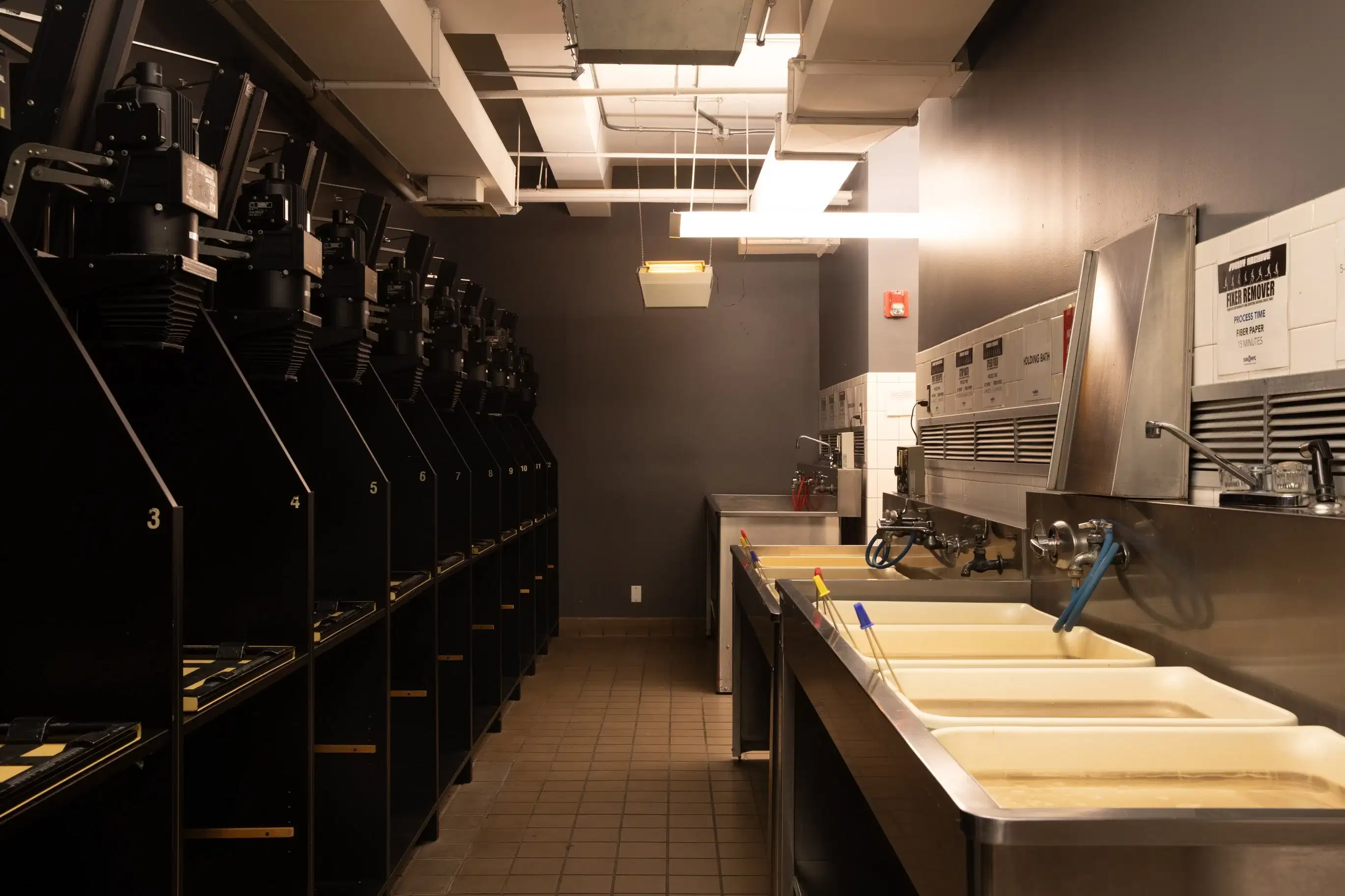 Dark room with a row of enlargers on one wall and and row of sinks with developing chemicals on the other wall
