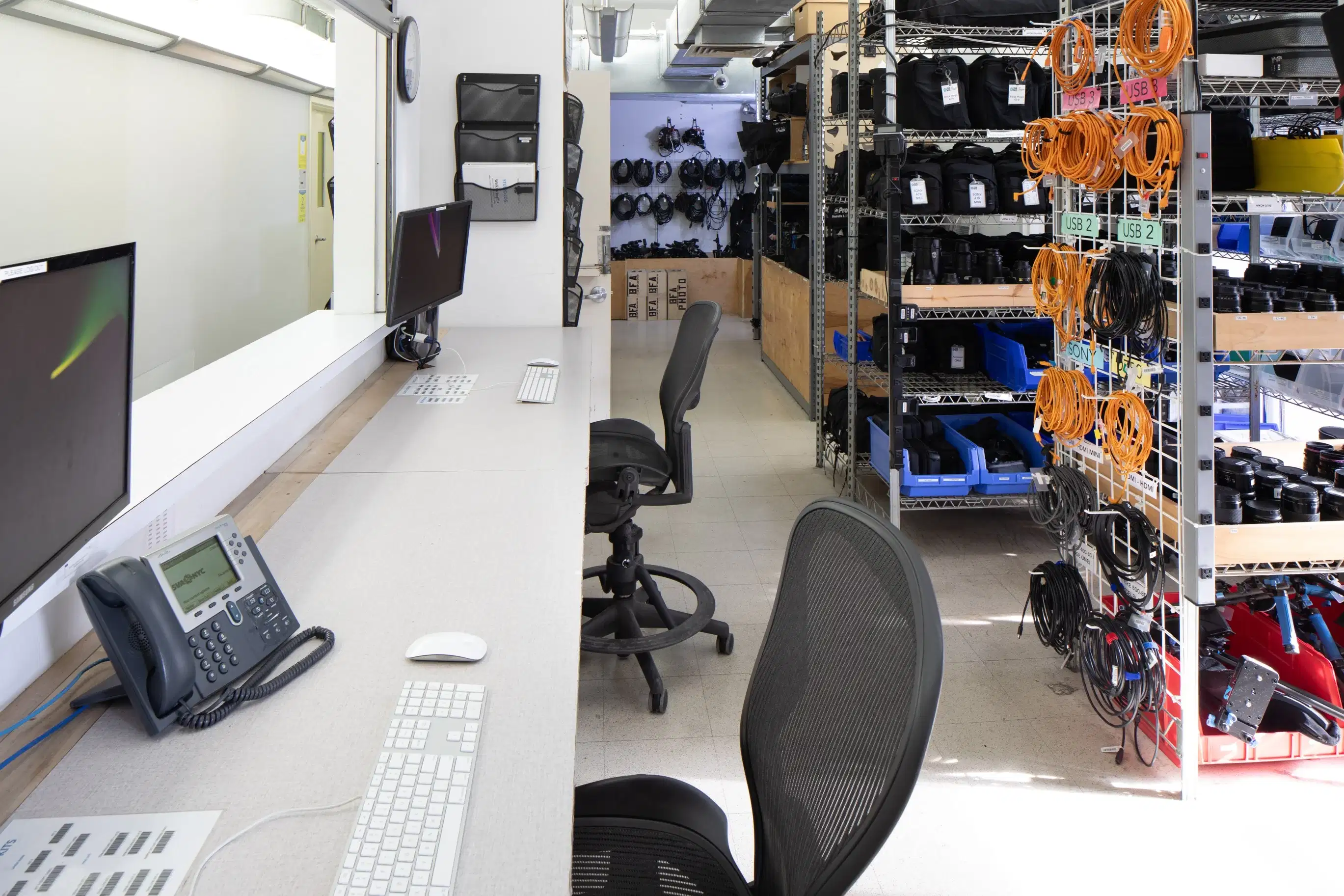 A long work station with two chairs and computers facing a window. Organized cameras and lenses on shelves behind the station.
