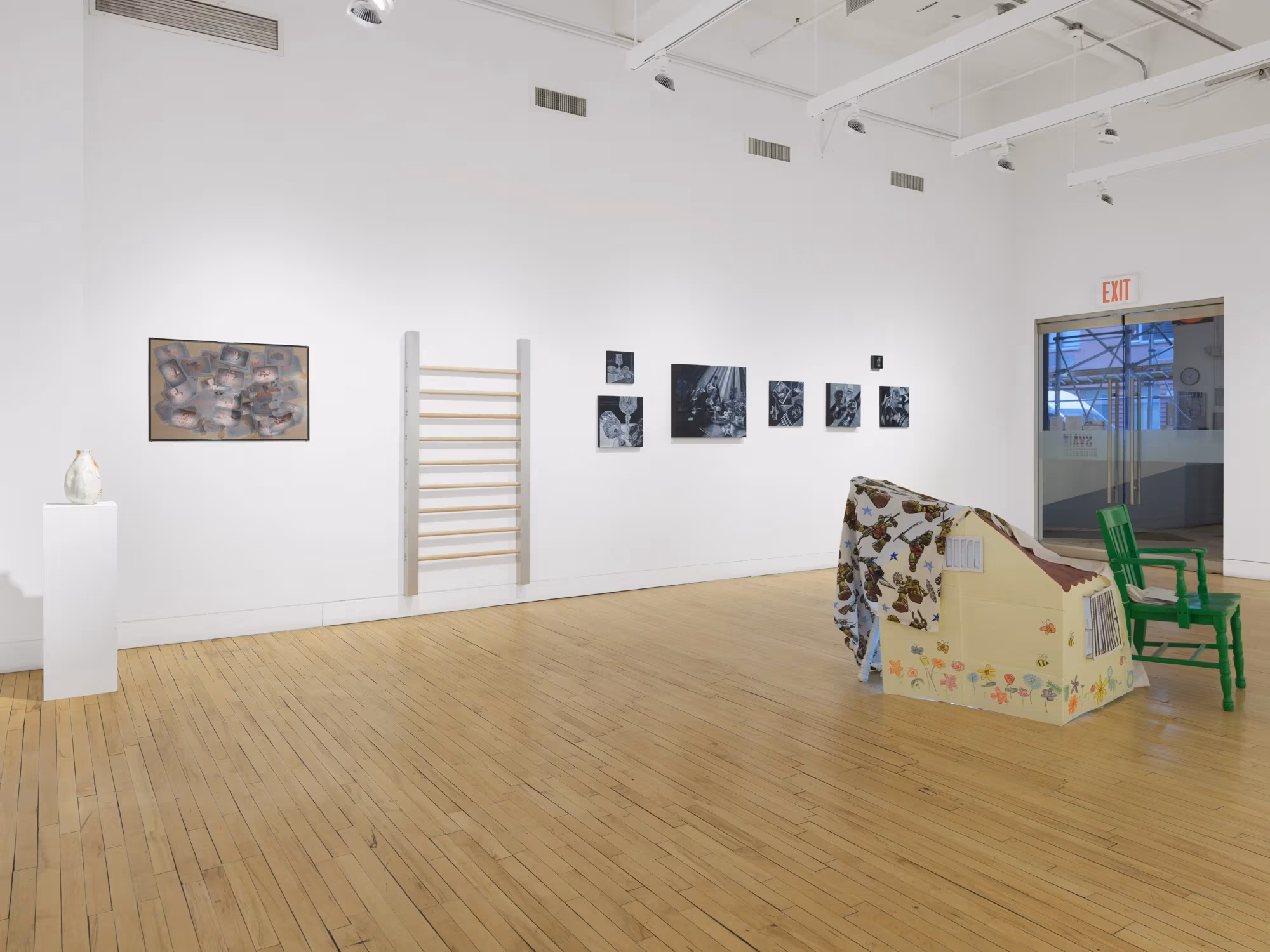 A wall of the gallery filled with several paintings and sculptural pieces, including one that resembles a ladder in the center of the wall.