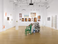 Each wall of the gallery is filled with several different artworks and paintings, and in the center is a sculptural piece containing a green chair.