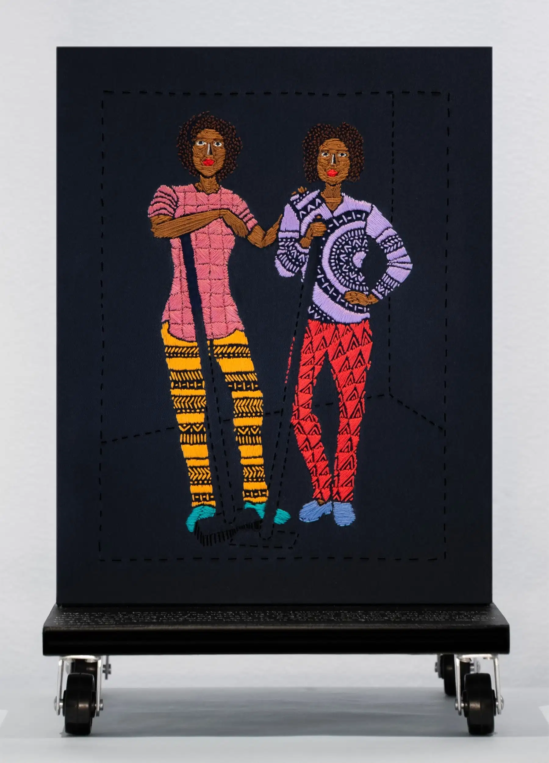 A black embroidered canvas with two figures of women standing and holding cleaning supplies.