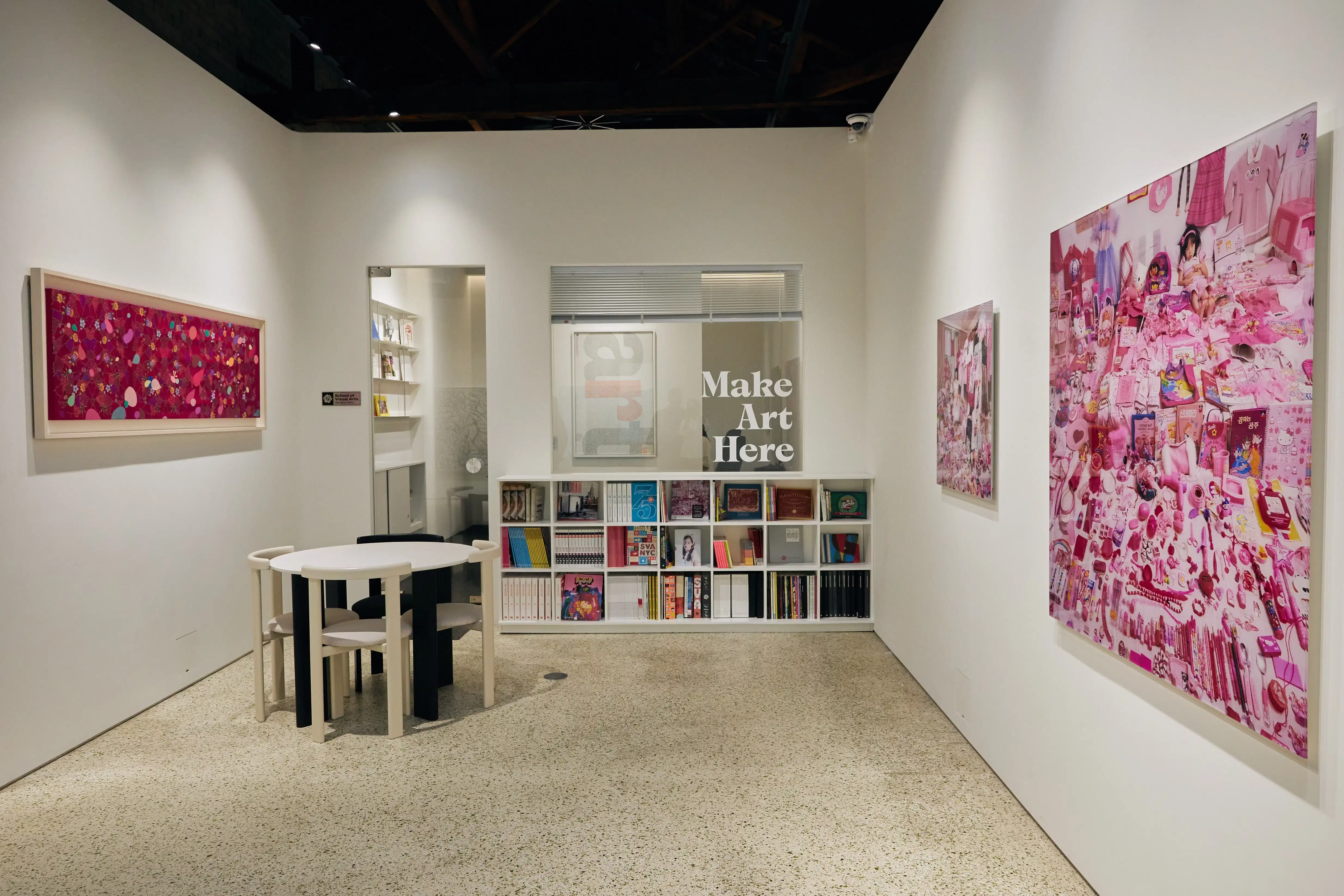 The image displays a photograph of a nook and office space within an art gallery space; a table and chairs are situated to the left, a bookshelf full of colorful art books in the middle and the words "Make Art Here" written on the glass of the office space. 