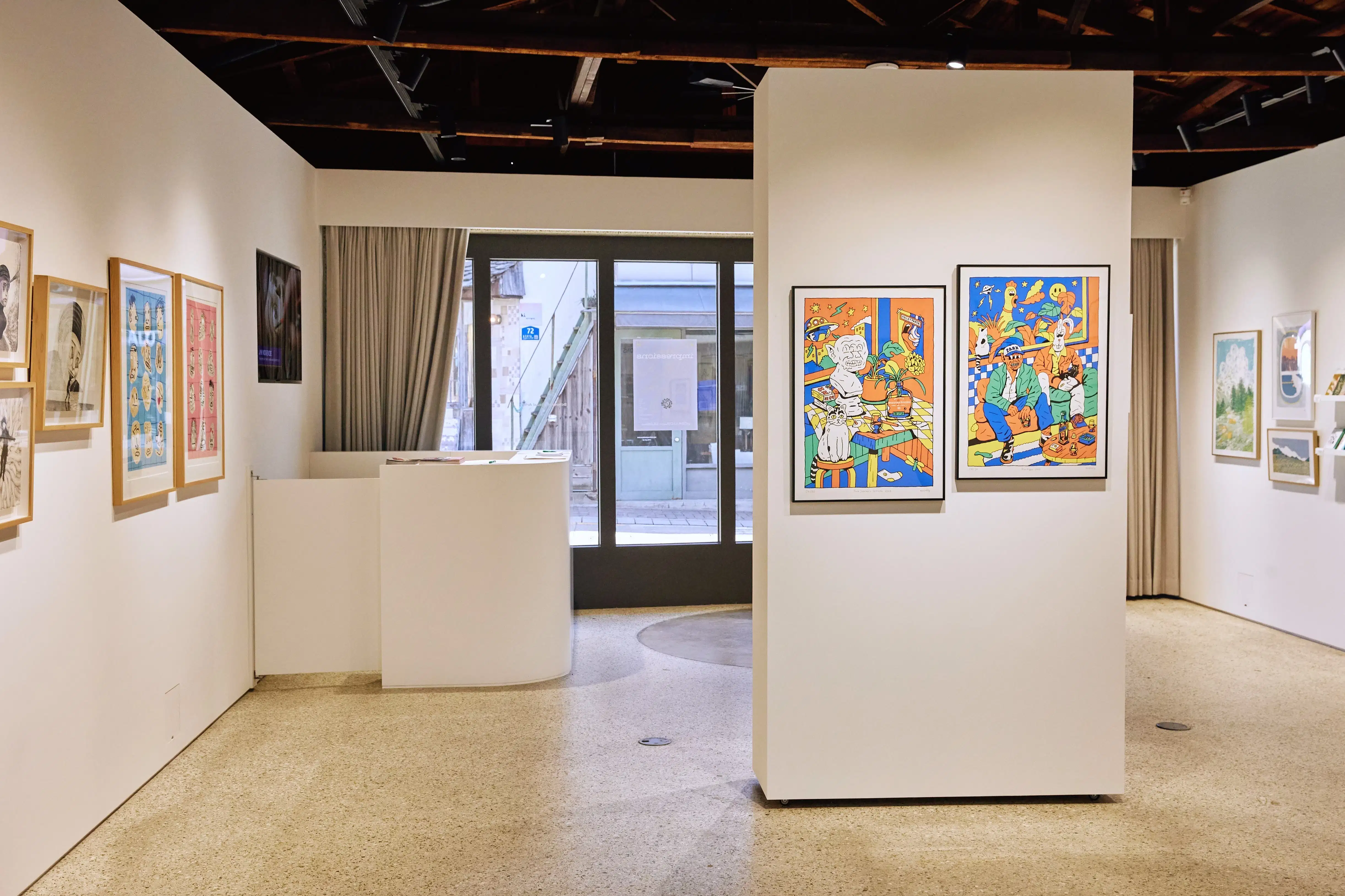 Gallery interior featuring colorful cartoon-style illustrations on a central wall, surrounded by framed drawings, portraits, and nature-themed artworks