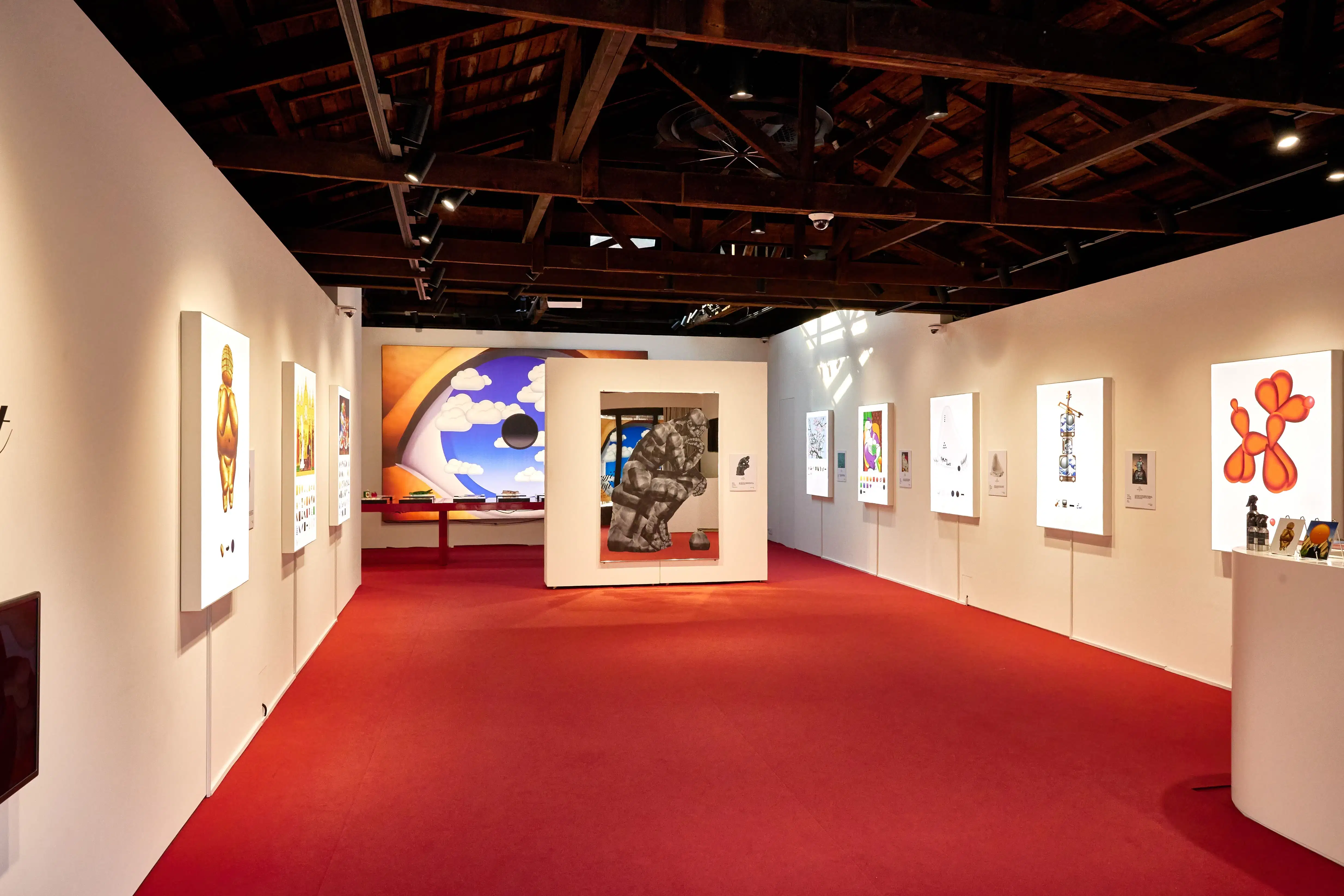Gallery view of a contemporary art show with pop and surrealist works: colorful balloon-like figures, a sky painting, and a central seated sculpture.