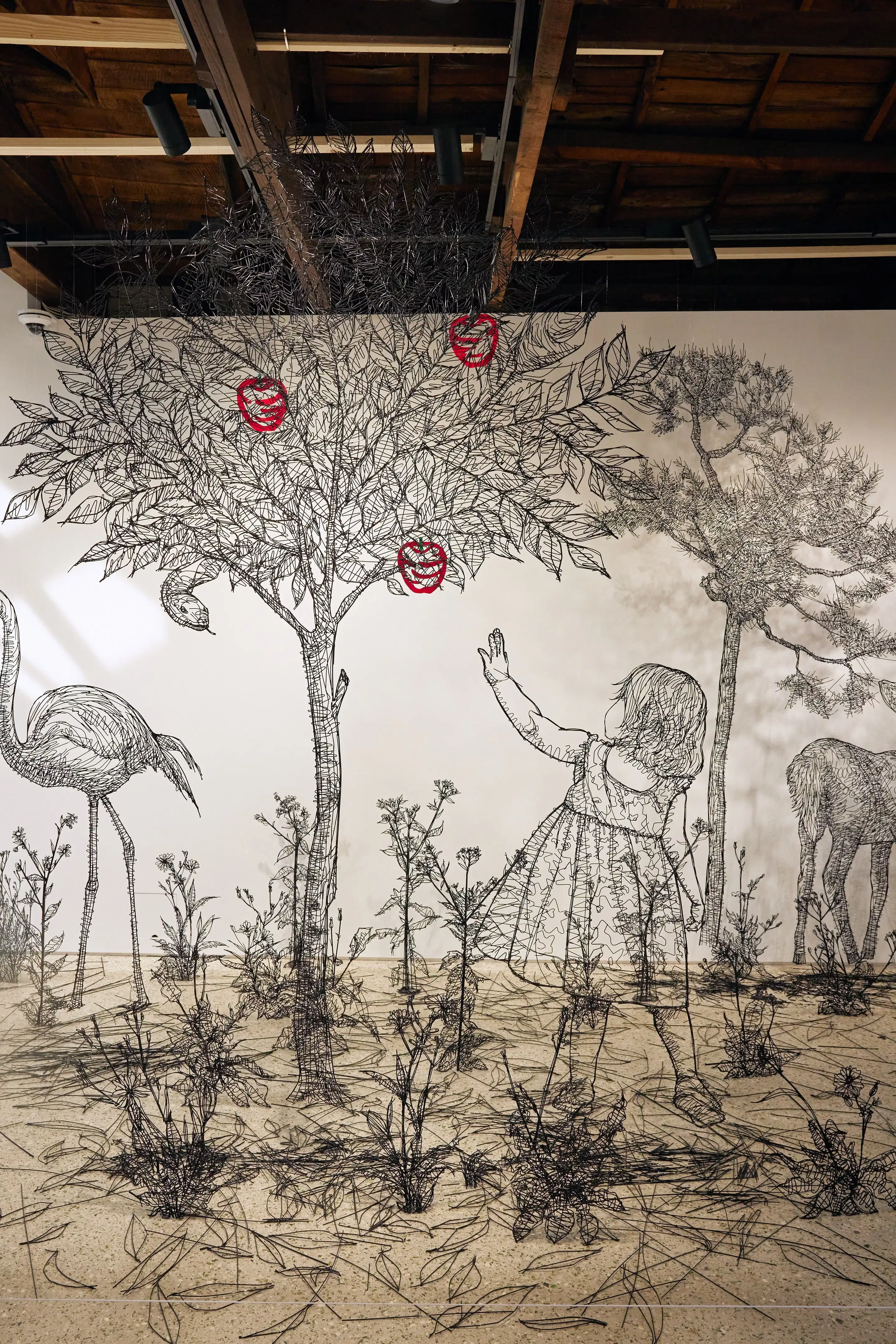 A three-dimensional installation on a white wall constructed with thin black plastic sticks that creates the illusion of a 2D drawing in three-dimensional space. Against a white background, this minimalist piece features sleek black linear elements depicting an apple tree surrounded by bushes, with a girl reaching for an apple on the right side.
