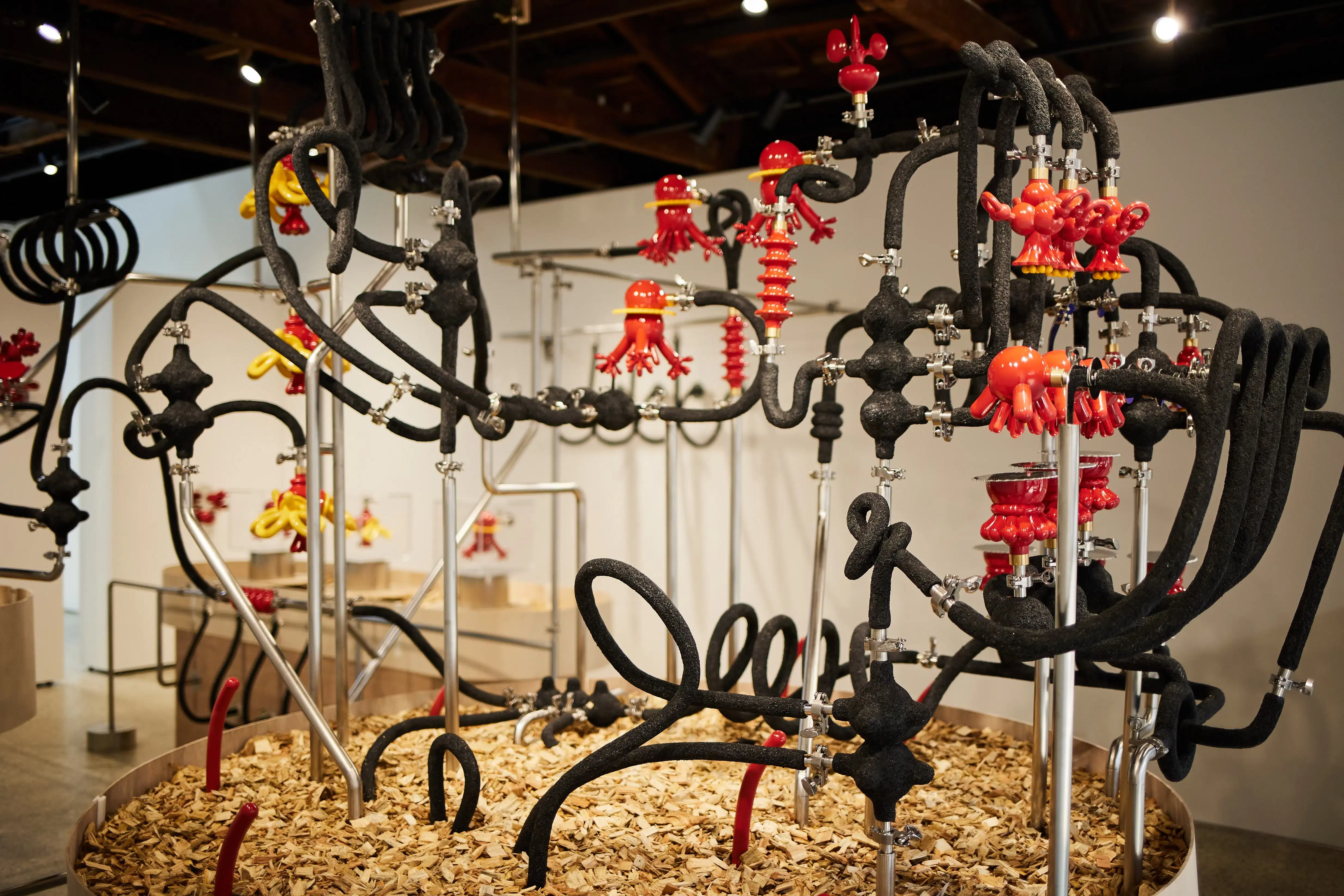 Complex sculptural installation featuring black foam-wrapped tubes and metal pipes twisting into loops and knots, with glossy red and yellow balloon-like glass figures resembling jellyfish and animals attached throughout. The structure is anchored in a circular base filled with wood chips, situated in a well-lit contemporary gallery.