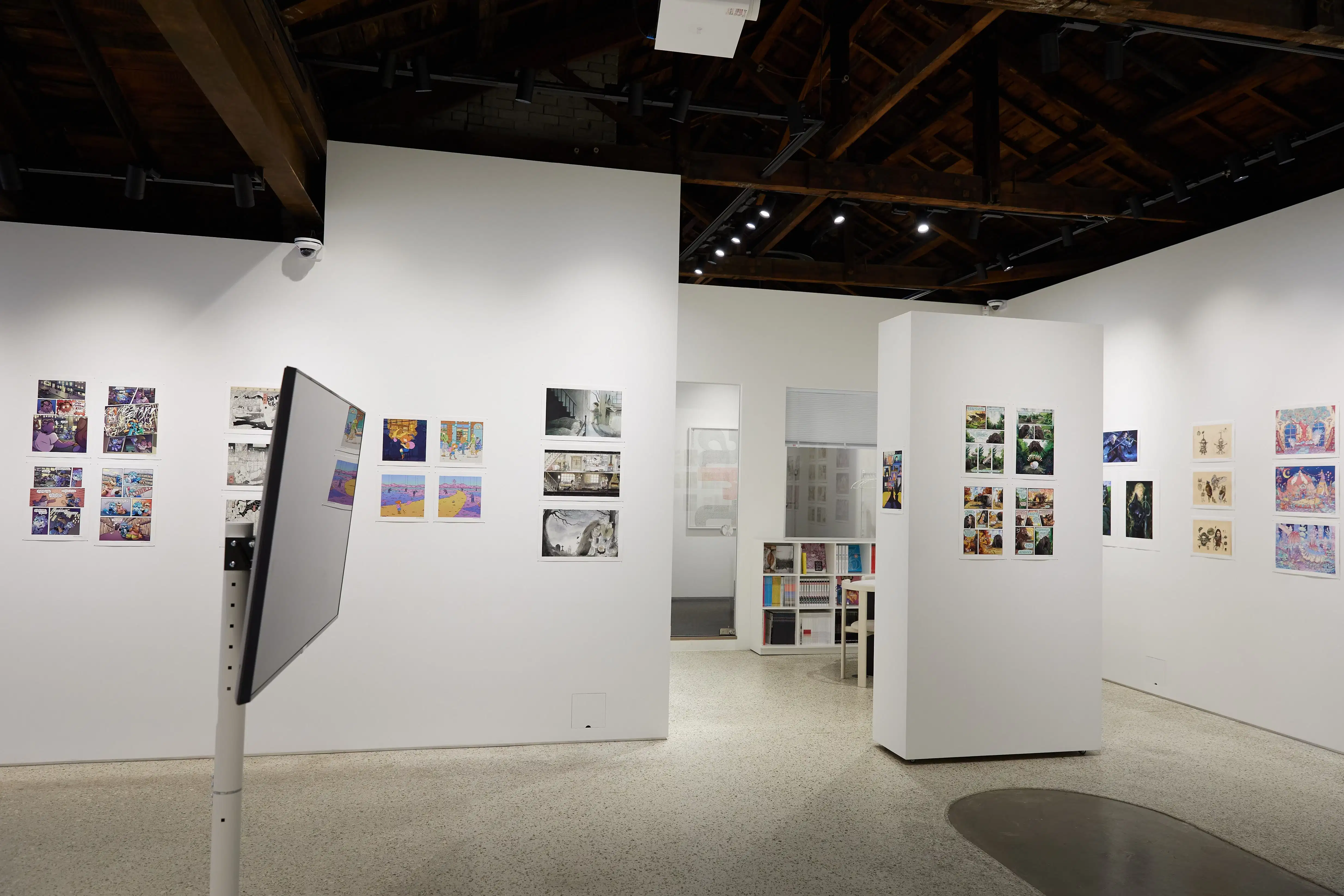 Gallery with comic and illustration artworks displayed on white walls and panels under an exposed wooden ceiling, with a standing monitor and bookshelves in the background.