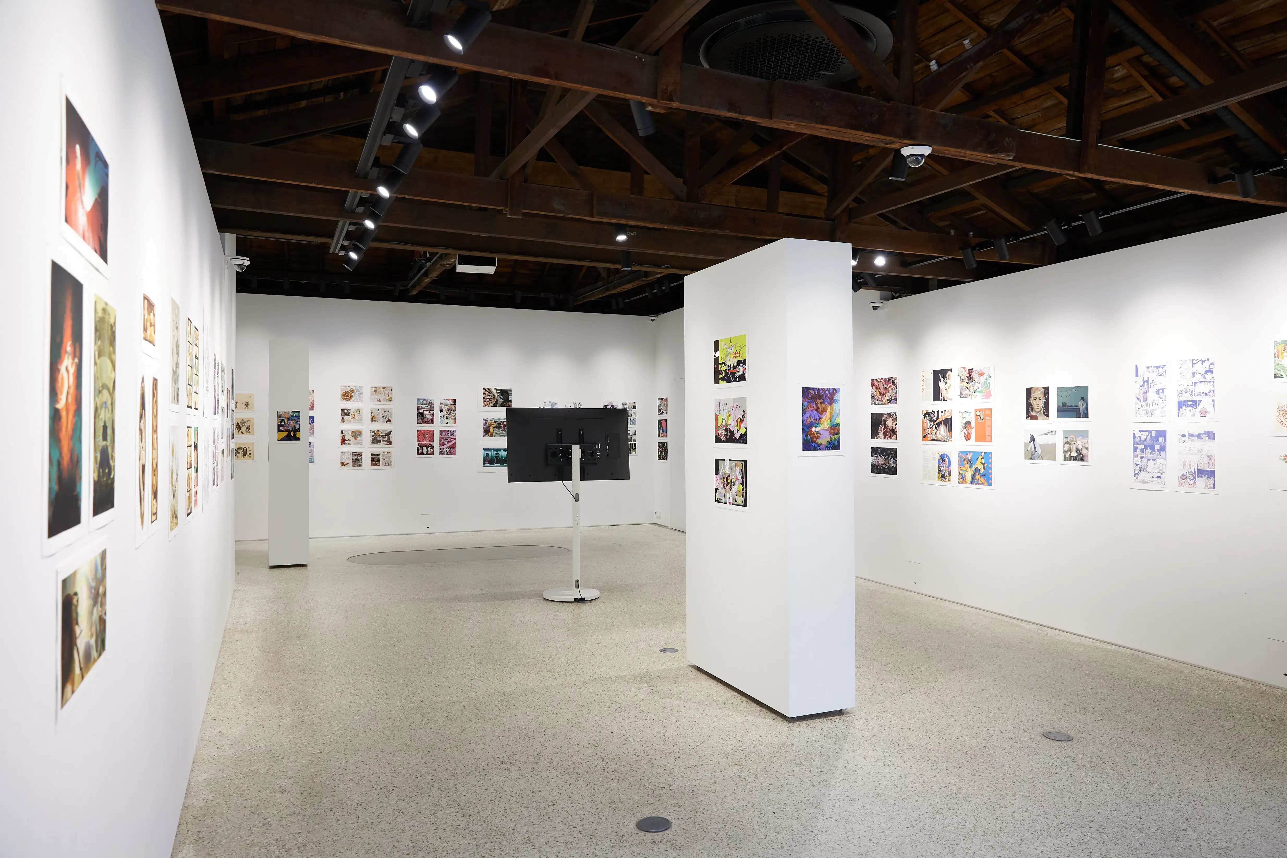 Spacious gallery interior with colorful comic and illustration artworks on white walls, a central monitor display, and an exposed wooden beam ceiling.