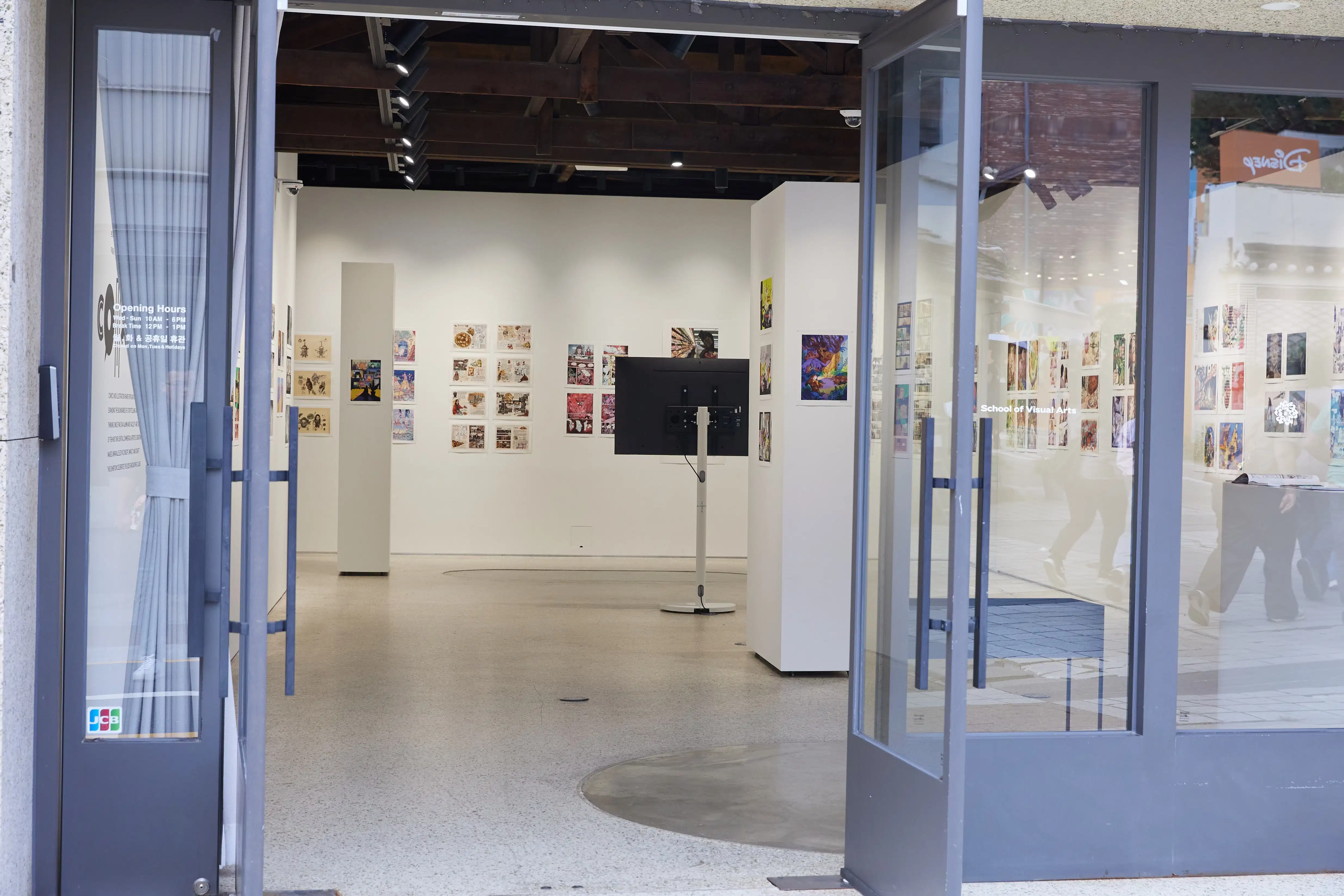 View through open glass doors into a bright gallery with comic and illustration artworks on white walls and a standing monitor at the center.