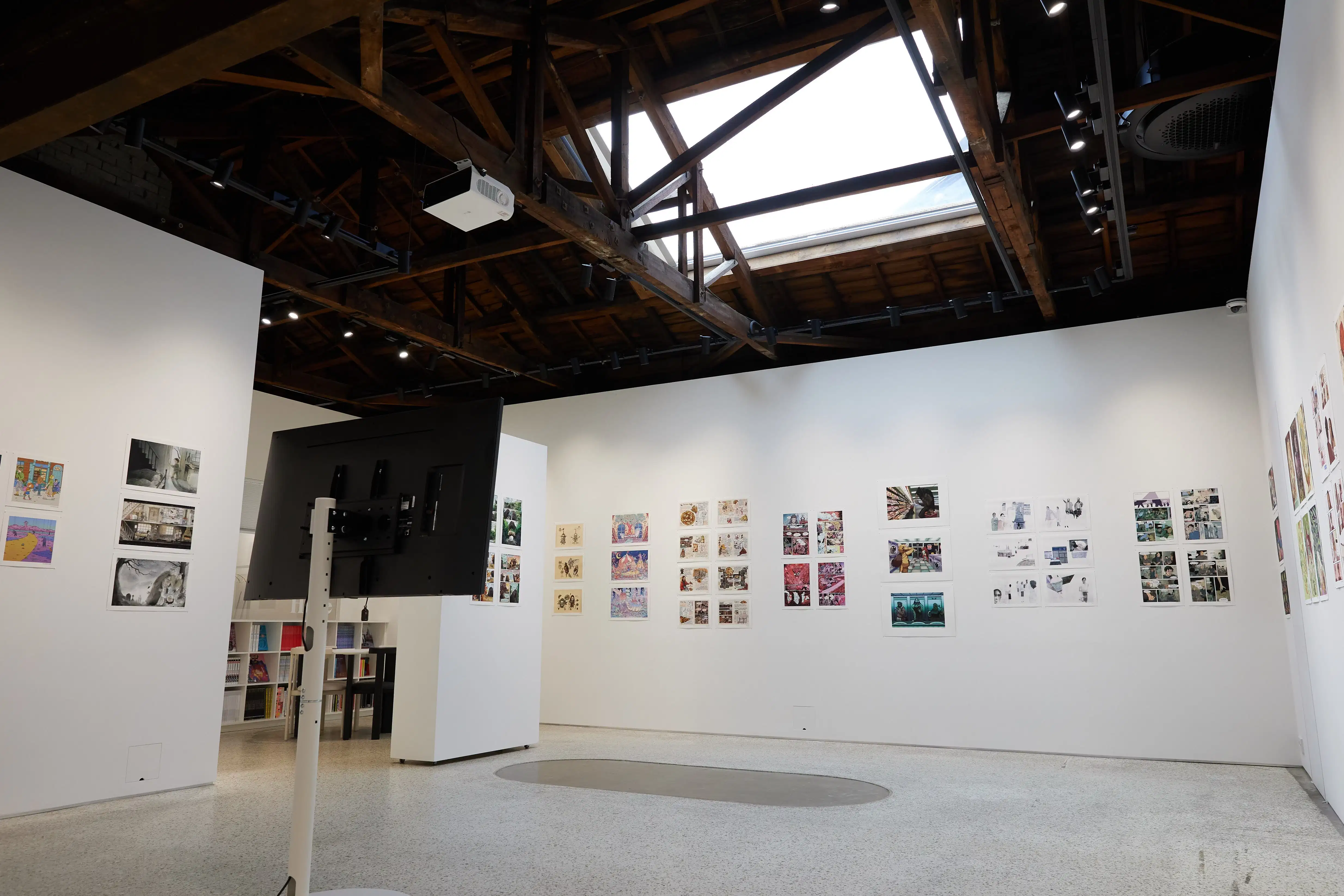 Gallery interior with colorful comic and illustration artworks on white walls, a standing monitor, and a wooden beam ceiling with a skylight above.