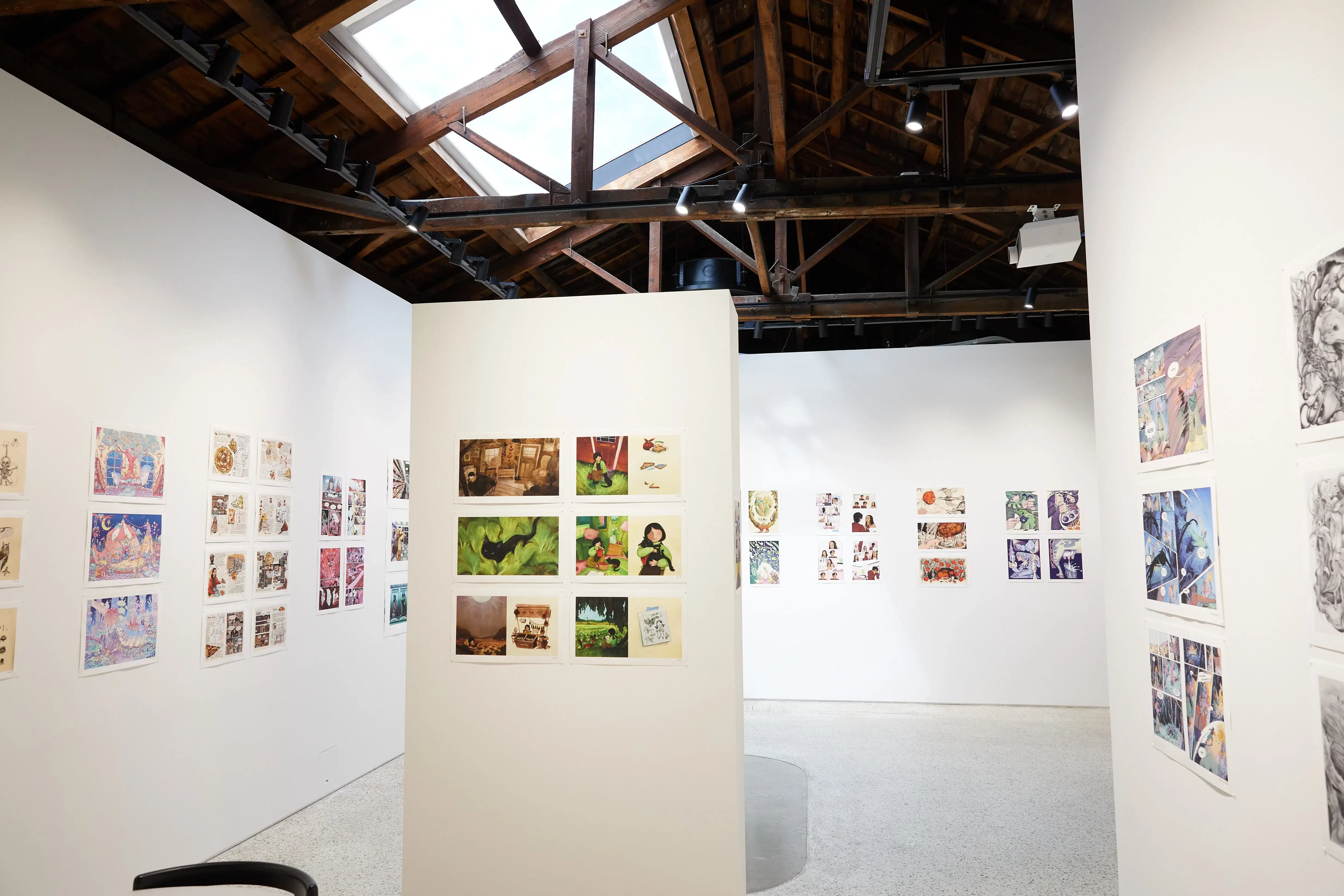 Gallery interior featuring colorful comic and illustration artworks on white walls under a wooden ceiling with exposed beams and a skylight.