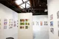 Gallery interior featuring colorful comic and illustration artworks on white walls under a wooden ceiling with exposed beams and a skylight.