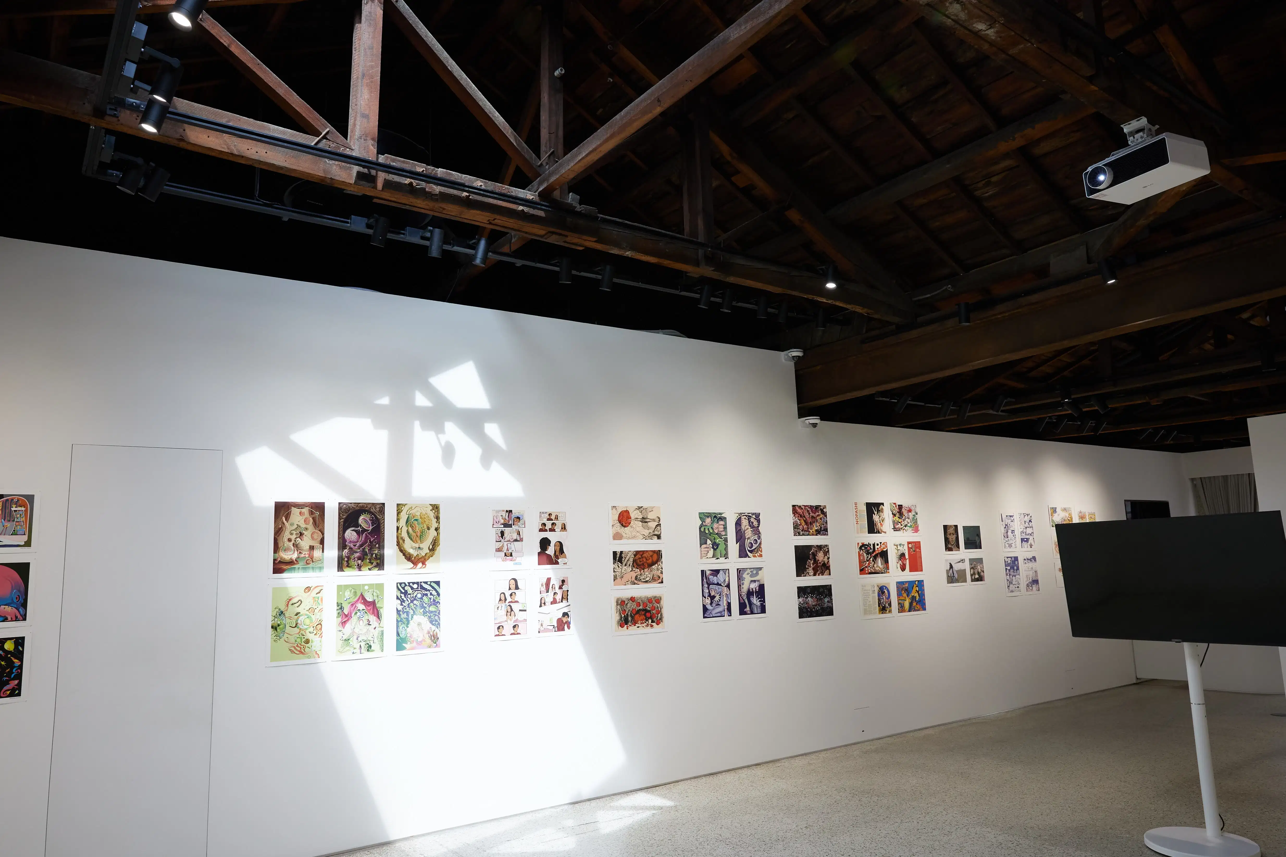 Gallery wall displaying colorful comic and illustration artworks under a wooden beam ceiling, with sunlight casting geometric shadows on the white wall.