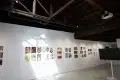 Gallery wall displaying colorful comic and illustration artworks under a wooden beam ceiling, with sunlight casting geometric shadows on the white wall.