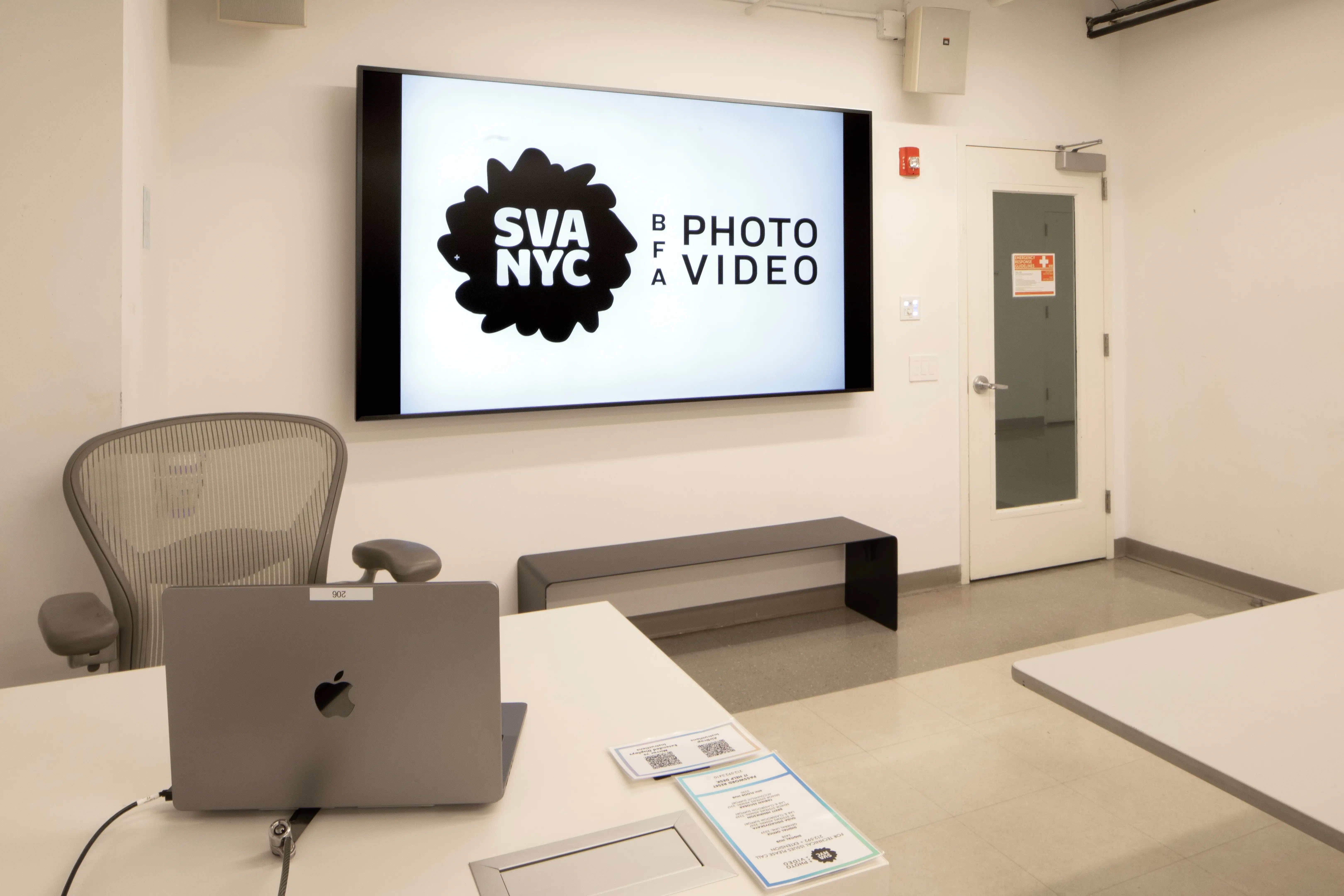 A clean classroom with a large monitor on the wall. The door, the end of a long table, and a smaller table with a laptop are all visible.