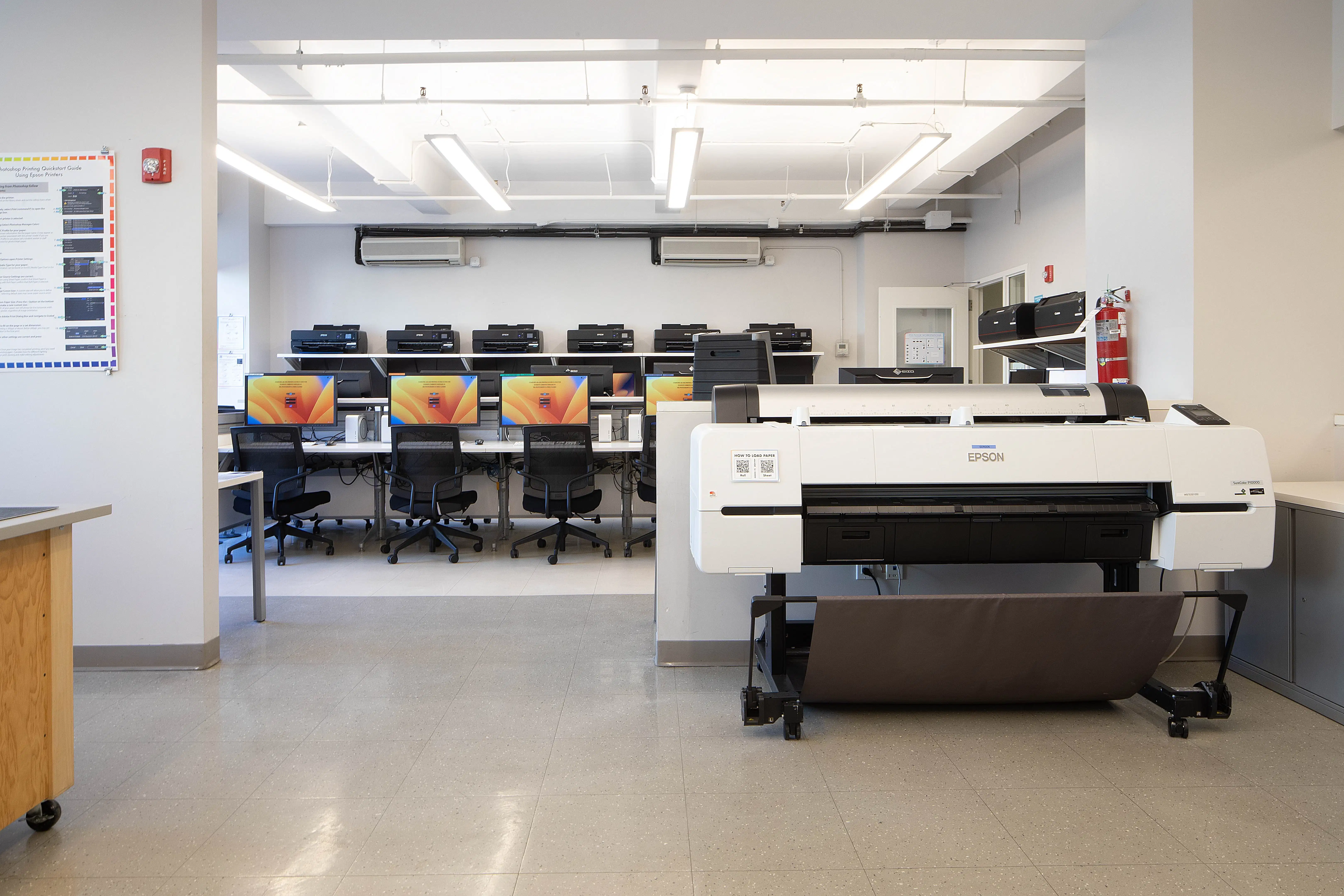 A large digital printer in front of a classroom of monitors and scanners.