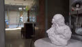 a  stone carving of a woman holding a bird sits on a marble table,  behind on the left we can see a figure out of focus through a glass door