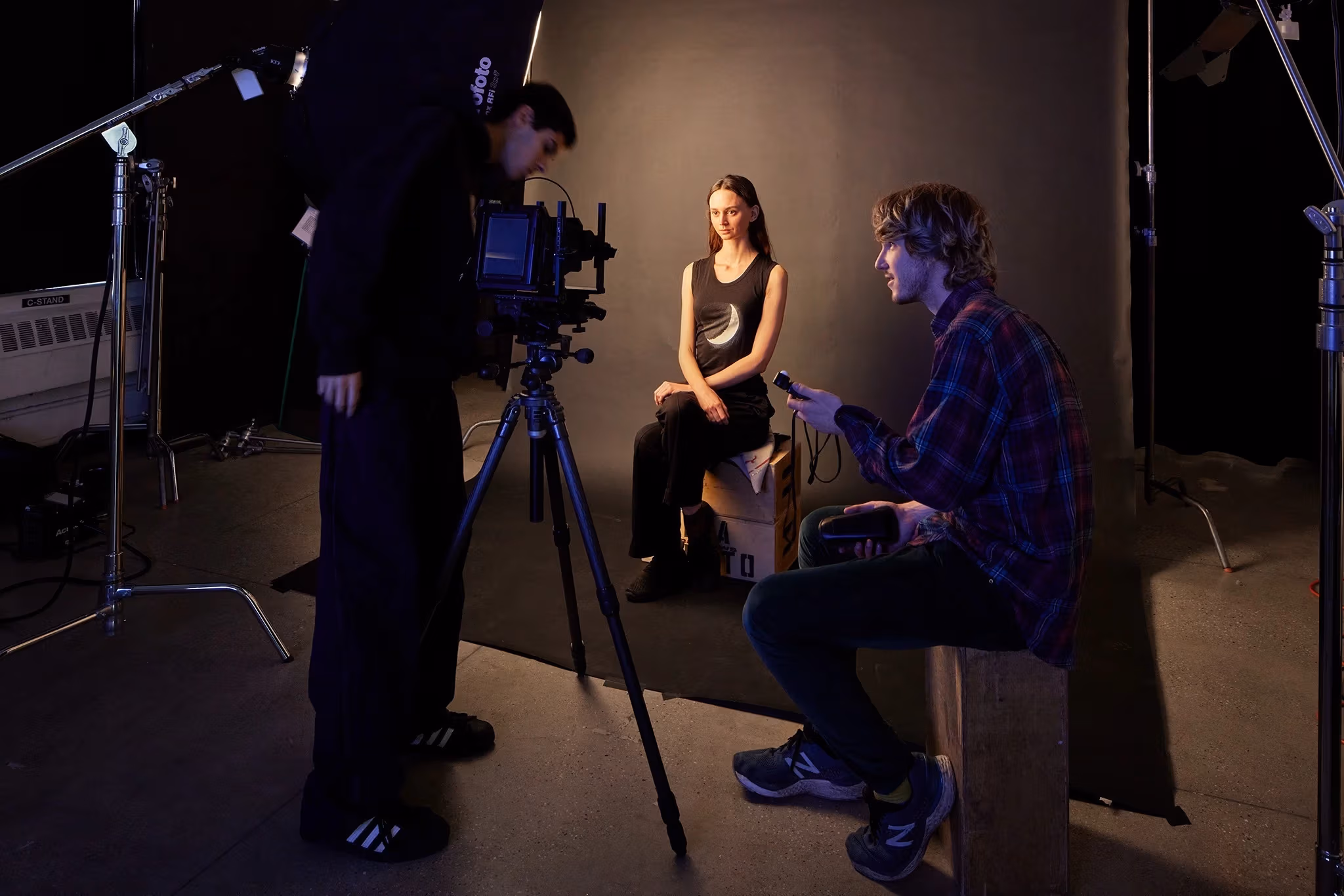 One person poses on an apple box while two others operate camera and sound equipment in a photo studio