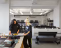 Two people look at digital prints on a table in front of a large digital printer